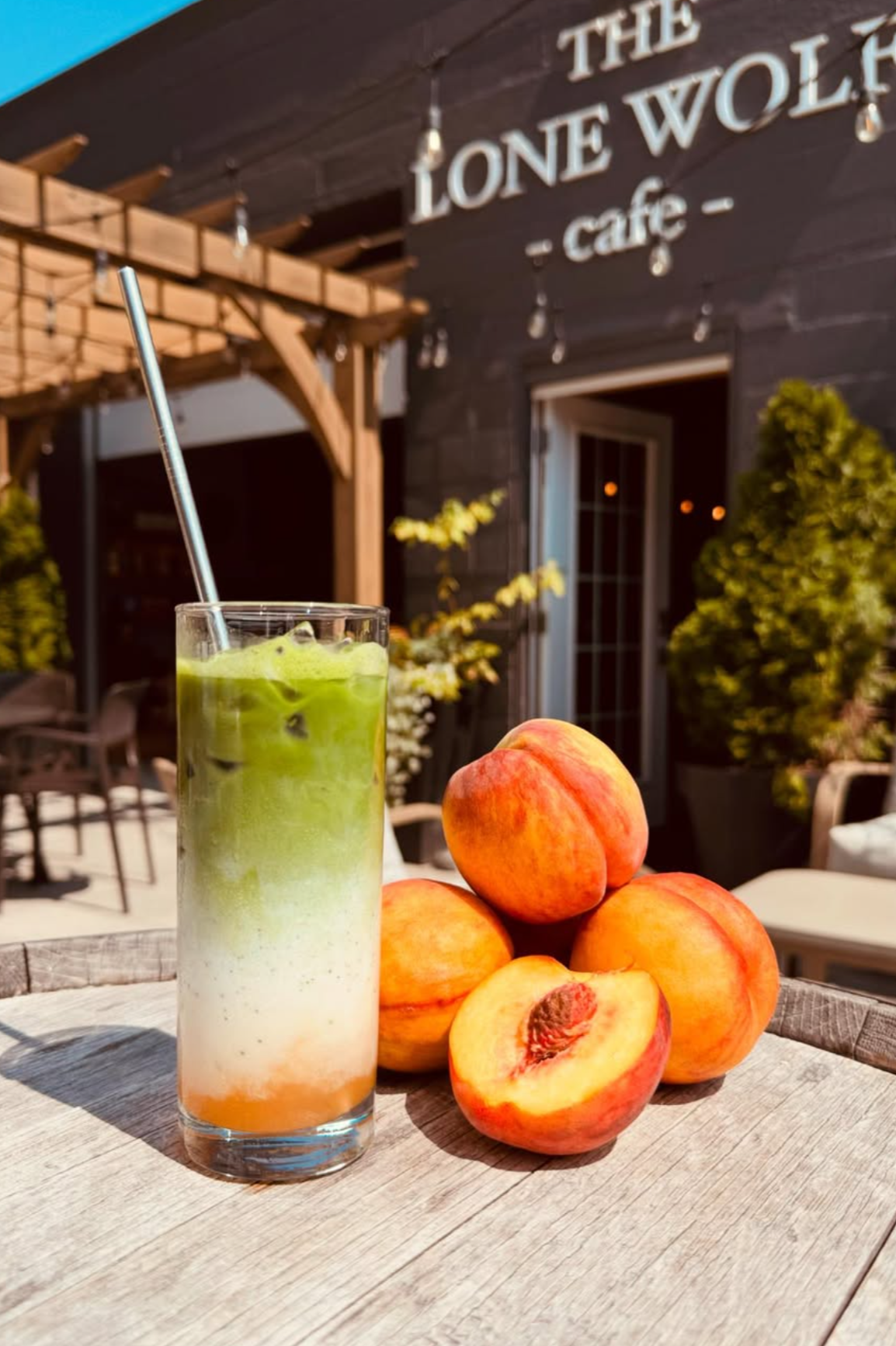 A colorful layered drink with a straw, five peaches, and one sliced open showing its seed, placed on a wooden outdoor table in front of a cafe called The Lone Wolf. The background shows a wooden balcony, green plants, and a sign reading 'The Lone Wol