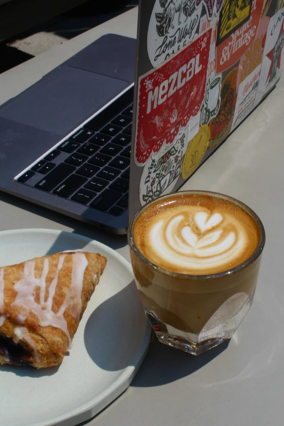 A coffee drink with latte art, a scone with icing, a laptop, and a sticker-covered box on a white table.