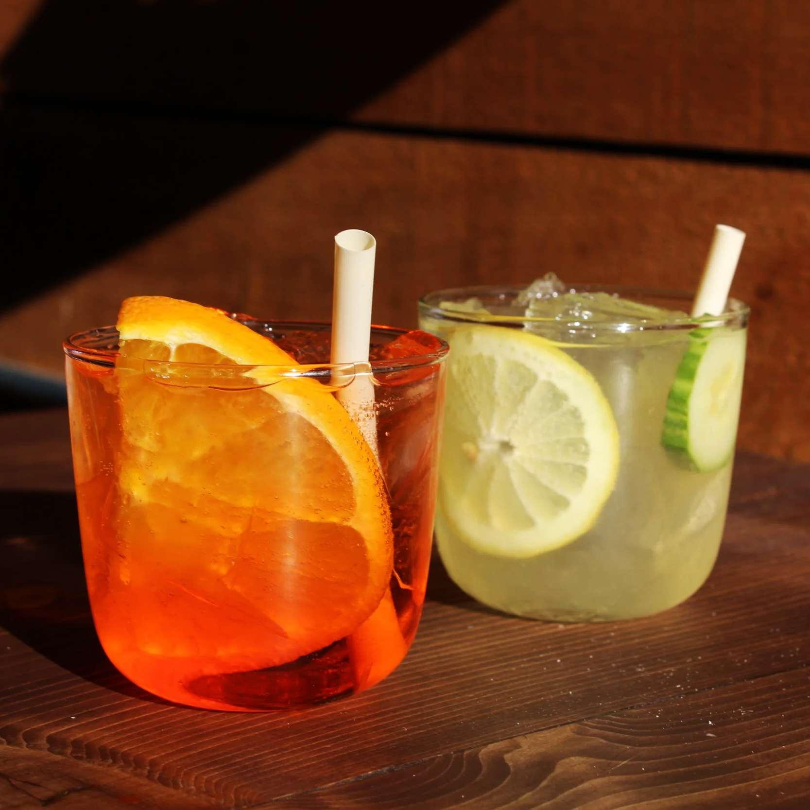 Close-up view of two cocktails on a wooden bar: one orange-colored with an orange slice, and one clear with lemon and cucumber slices, each with a straw.