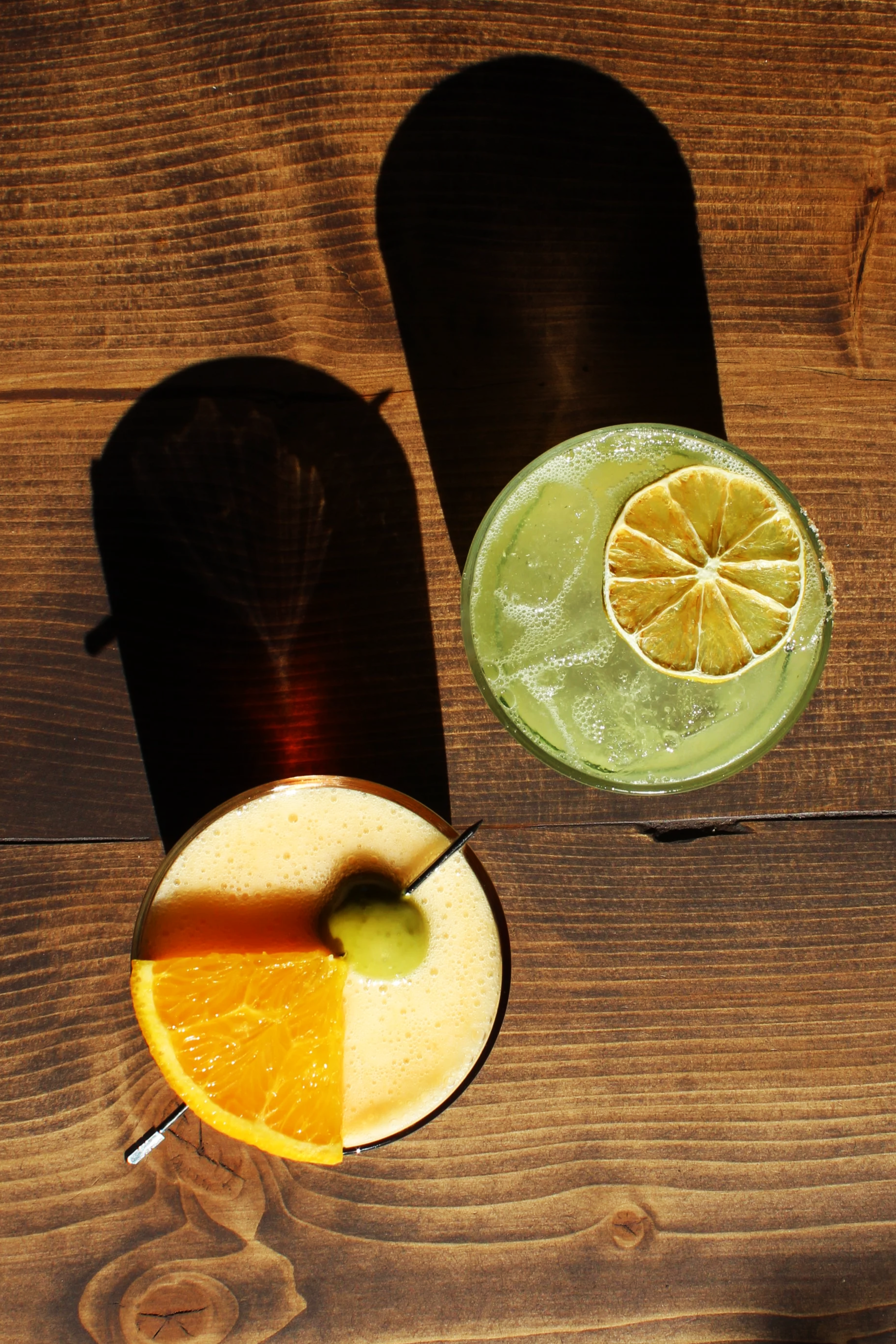 Two colorful cocktails on a wooden table, one with an orange slice and the other with lemon slices, each casting shadows