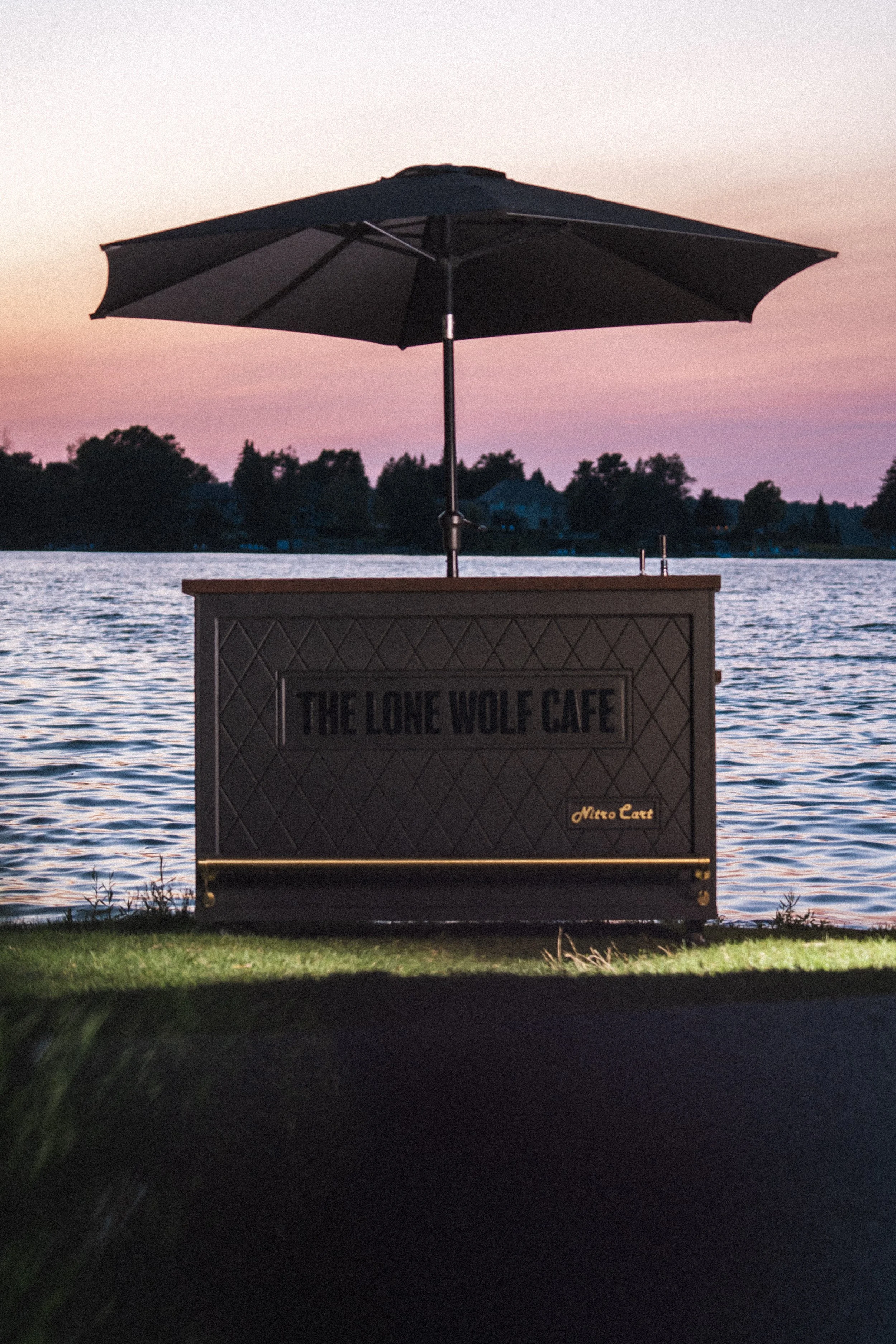 A lakeside scene at dusk with a black umbrella over a wooden sign that reads, "The Lone Wolf Cafe," and some grass in the foreground. The water reflects the pink and purple sky, with silhouettes of trees and houses in the distance.