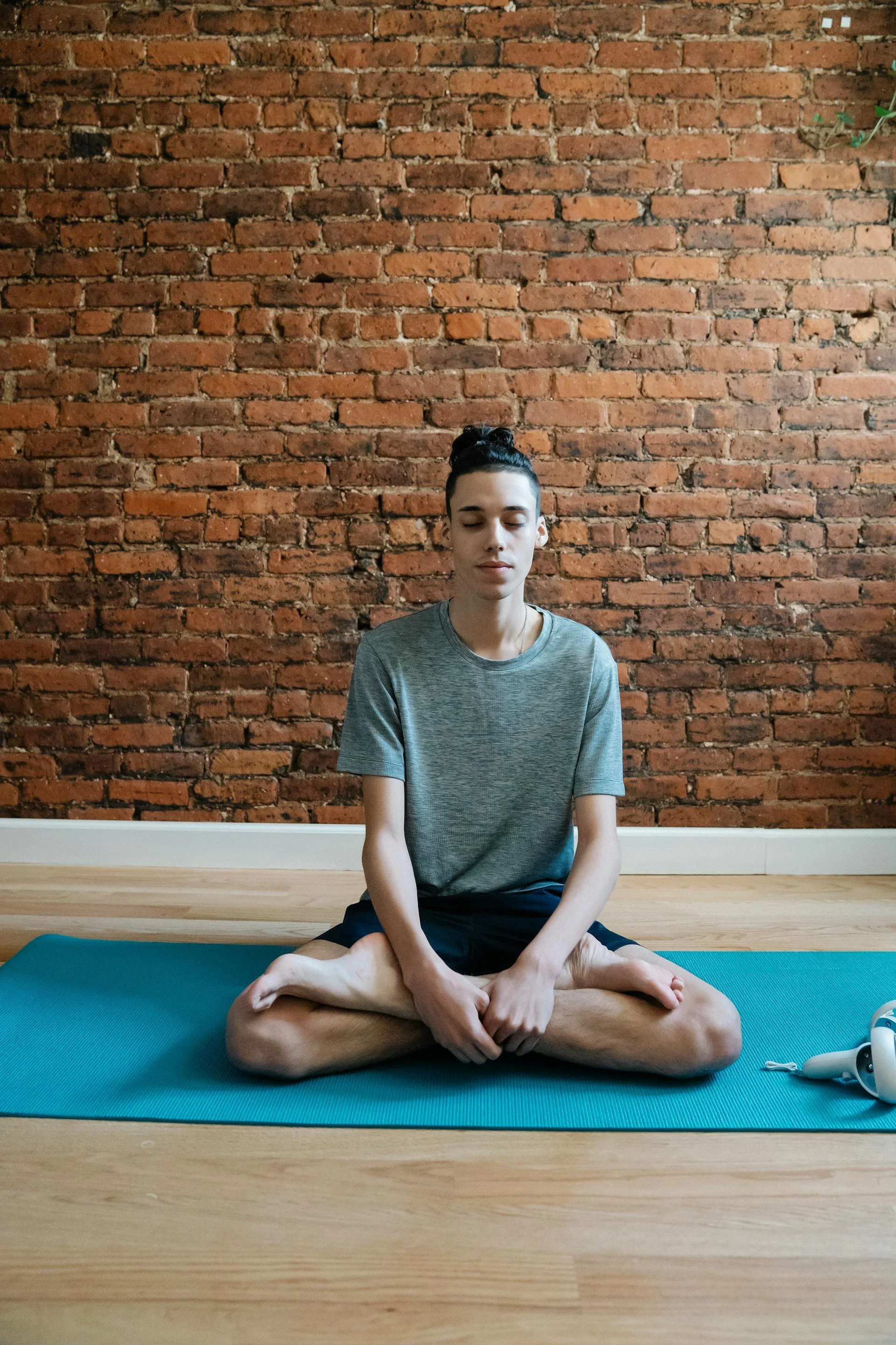 A person practicing yoga or meditation in a seated cross-legged position on a blue yoga mat indoors, with a brick wall in the background.