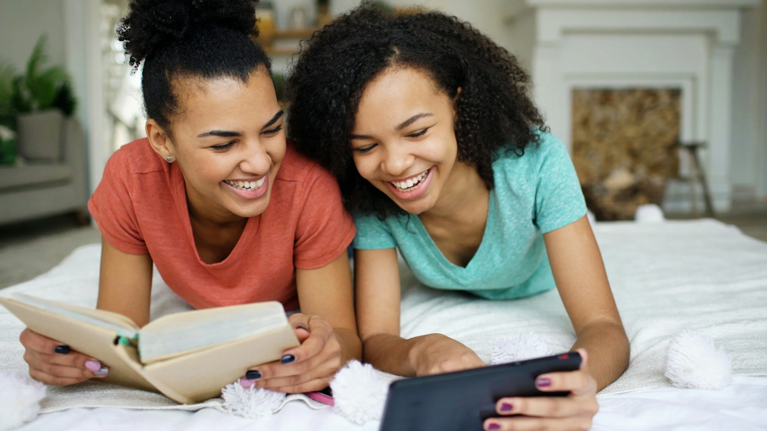 Two smiling women are lying on a bed, looking at a smartphone and a book together in a bright, cozy room with a fireplace and plants in the background.