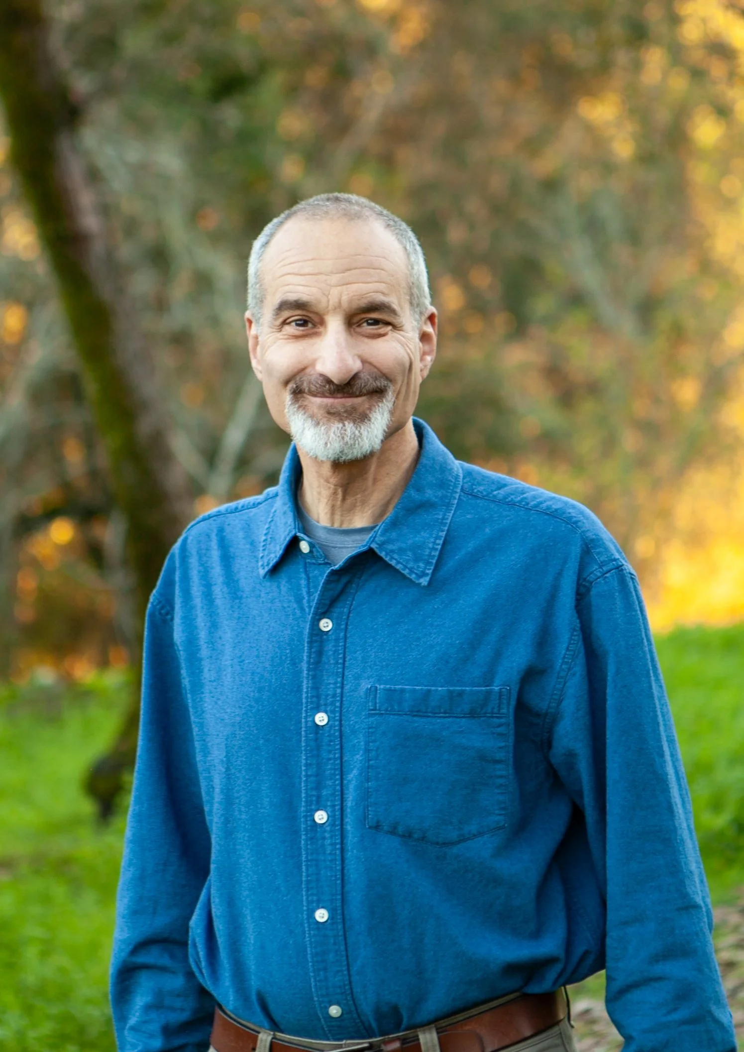 Portrait of a middle-aged man with a beard and short gray hair, smiling, standing outdoors in front of trees with autumn-colored leaves. He is wearing a blue button-up shirt and brown belt.