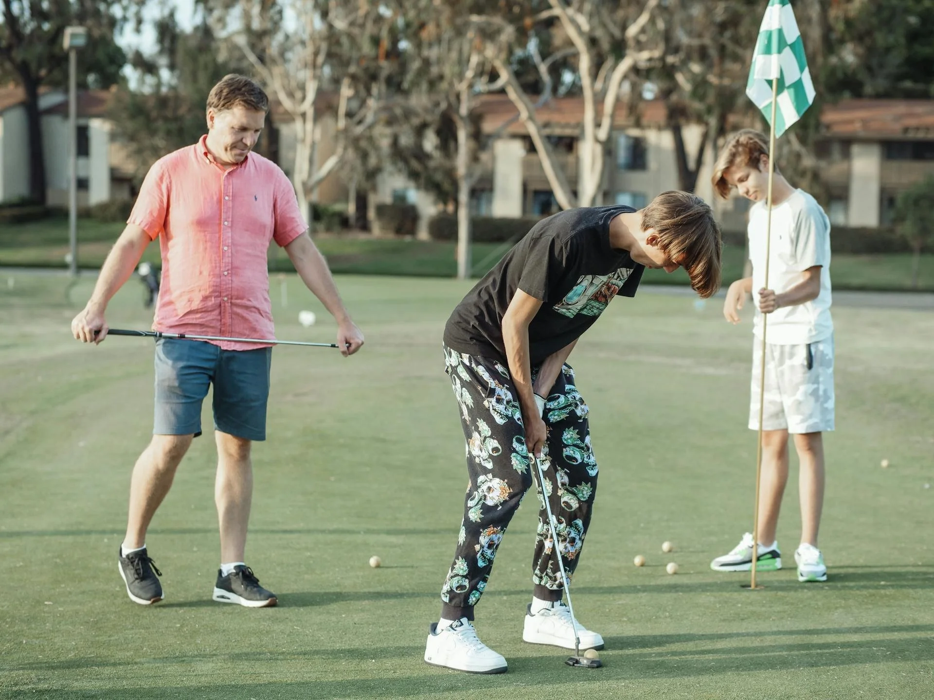 Four young men playing mini golf outdoors on a grassy course with a residential neighborhood and trees in the background. Two of them are preparing to putt the ball, while another stands with a golf club and a fourth holds a golf flag.