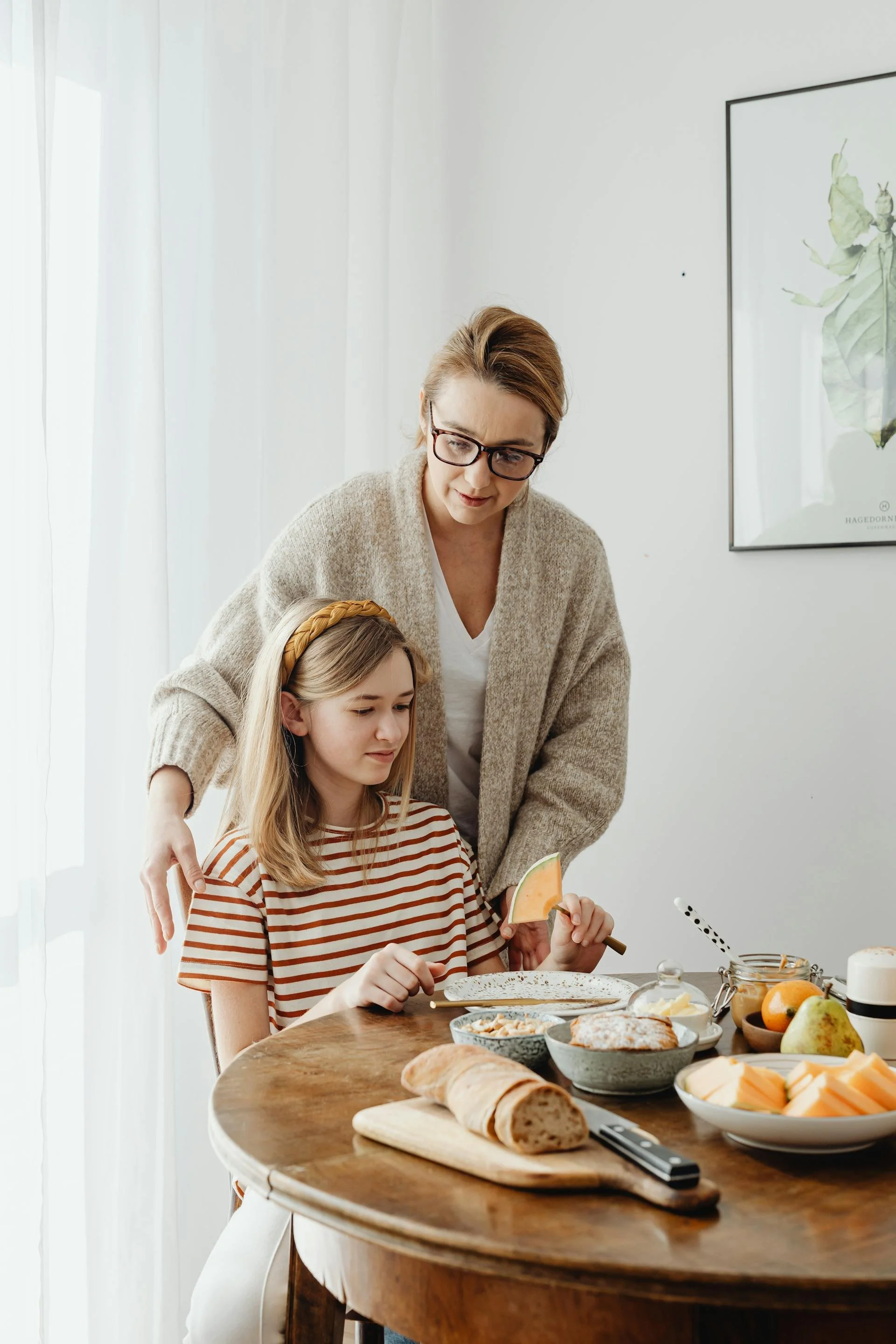 A woman and a girl with blonde hair are at a wooden dining table, preparing to eat a meal. The woman is standing, wearing glasses and a beige sweater, while the girl is sitting, wearing a red and white striped shirt. The table is filled with food, including sliced bread, cheeses, fruits, and spreads. The background features white curtains and a framed picture of butterflies on the wall.