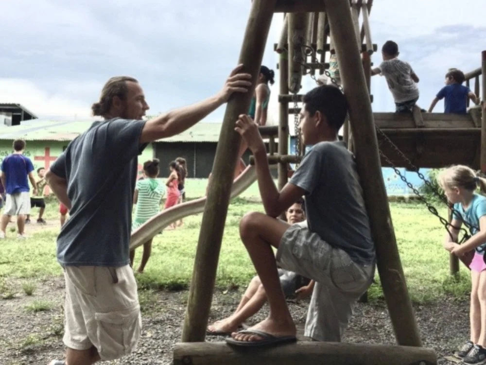 A man talking to a young boy sitting on a swing at a playground during daytime, with other children playing on the playground and around. The setting appears to be outdoors with a cloudy sky.