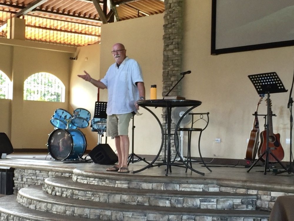 A man with a beard and glasses is standing on a stage with musical instruments, including drums and guitars, in the background. He is wearing a white shirt and shorts and appears to be speaking or presenting.