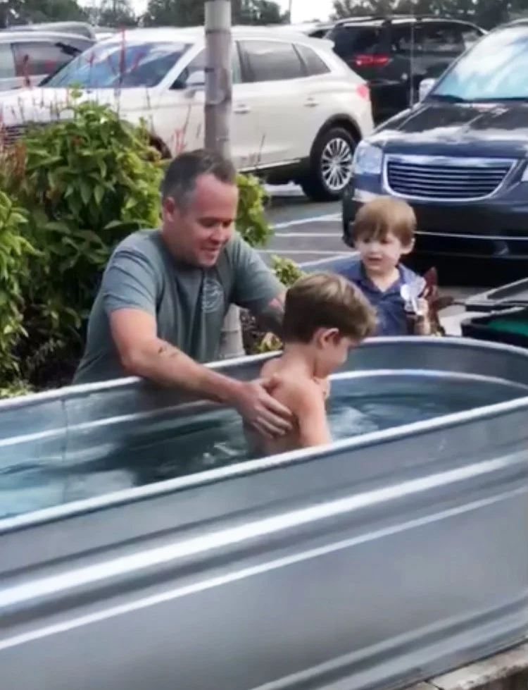 A man assists a young boy in a metal, rectangular baptismal tub filled with water outside in a parking lot, with another young boy observing nearby.