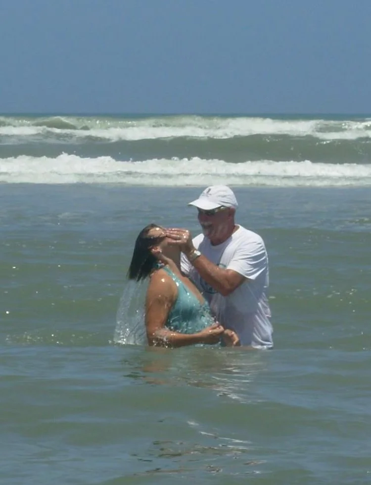 An older man and a woman are standing in the ocean, where the man appears to be performing a baptism.