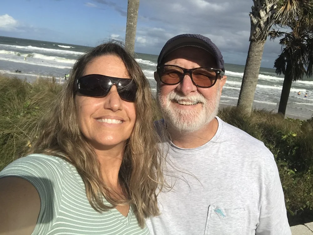 A smiling woman and man taking a selfie at the beach with ocean waves, palm trees, and a cloudy sky in the background.