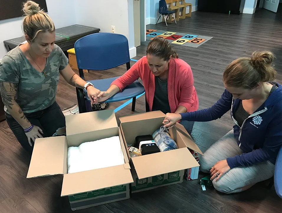 Three women are sitting and kneeling around open boxes filled with various items, possibly packing or organizing them in a room with wood flooring and chairs in the background.