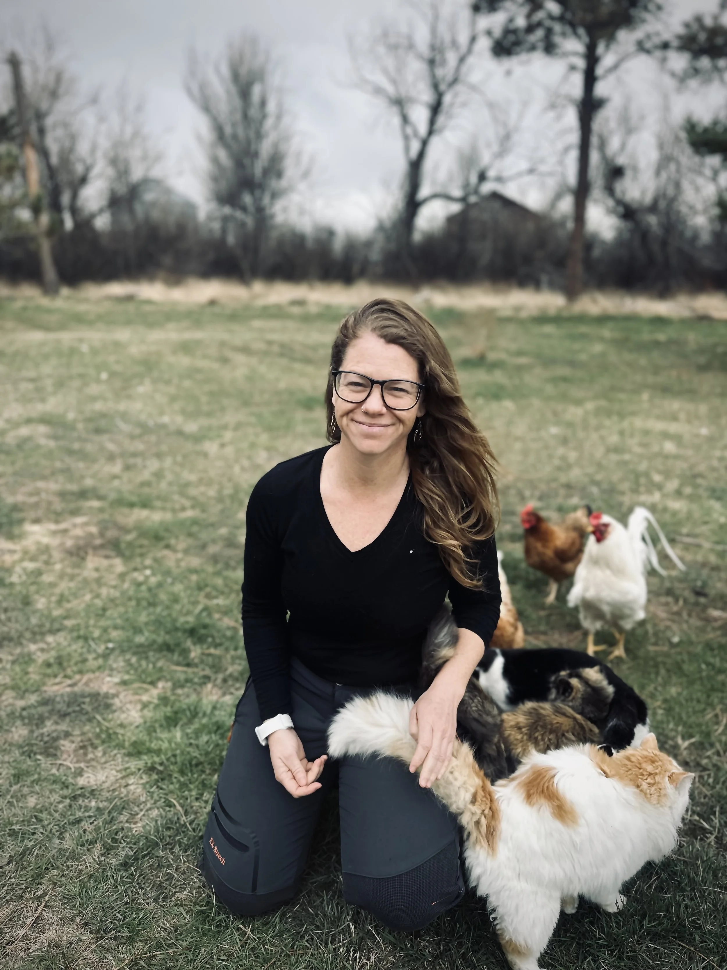 A woman with glasses in a black long-sleeve shirt and outdoor pants kneels on grass, smiling, surrounded by chickens and several cats outdoors with trees in the background.