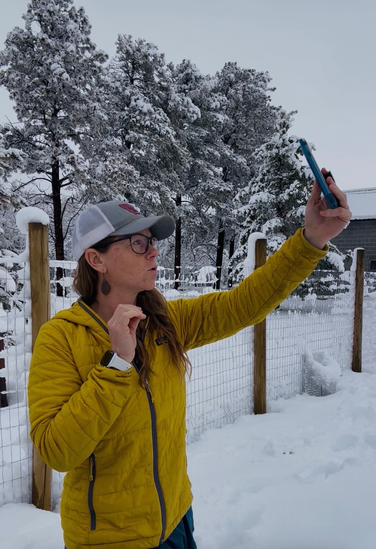A woman with long hair, glasses, and earrings taking a selfie in a snow-covered outdoor setting. She is wearing a yellow jacket and a gray cap, with a smartwatch on her wrist. There are snow-covered trees, a fence, and a building in the background.