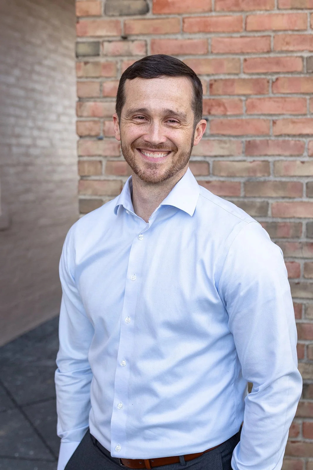 Anchor Point coach with short dark hair and a beard, wearing a light blue button-up shirt, standing in front of a brick wall.