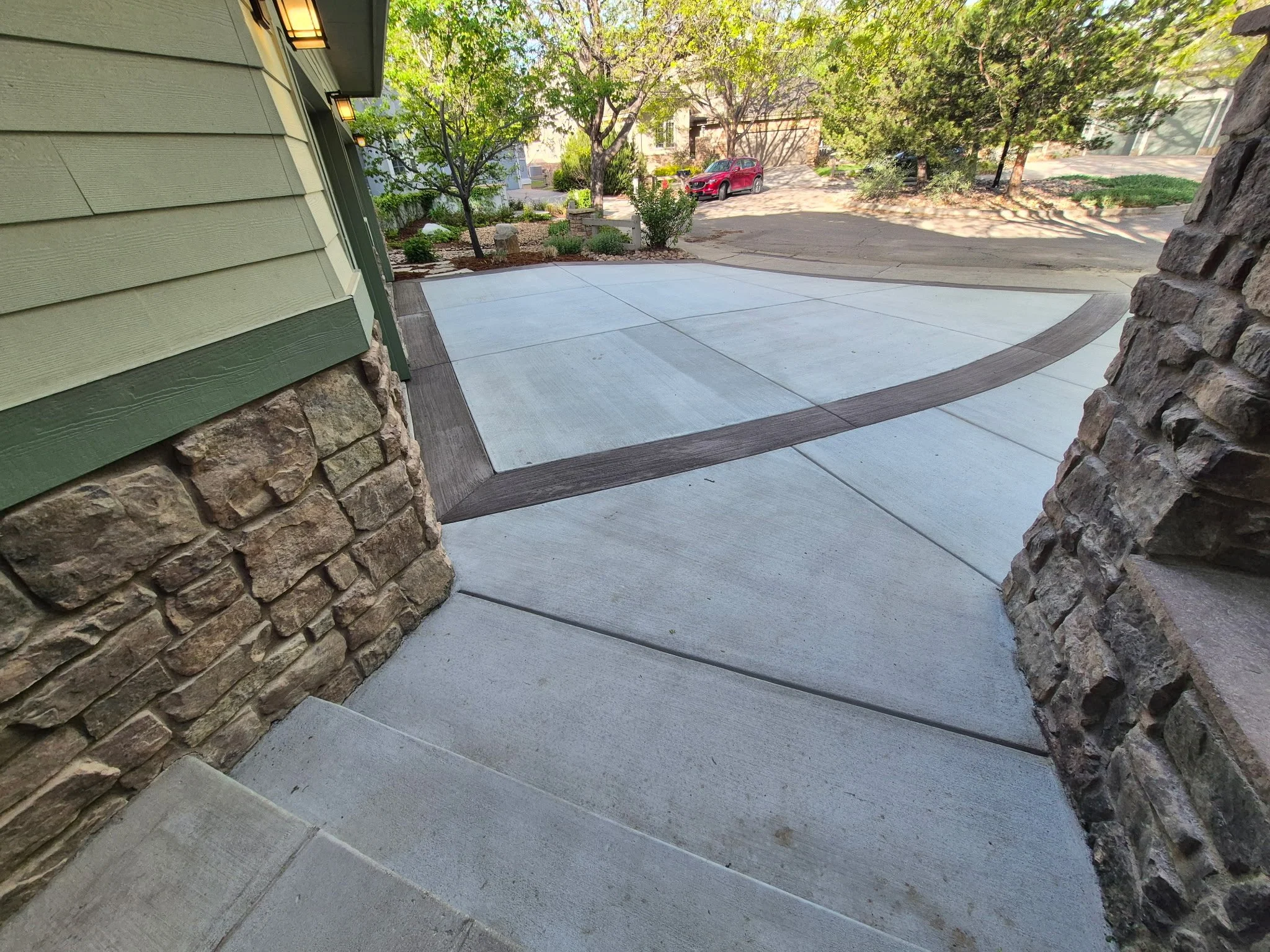 View of a concrete driveway with a decorative wood inlay pattern, bordered by stone walls and a green house with siding.