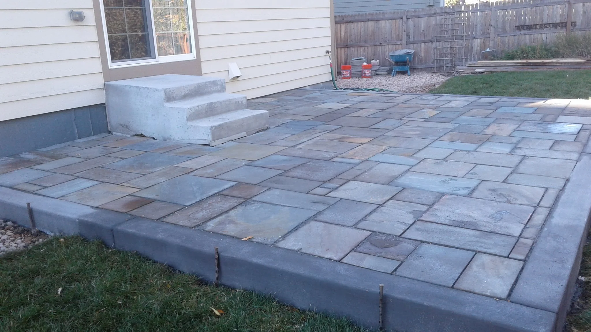 Newly completed concrete steps and stone patio outside a house with beige siding and a fenced backyard.