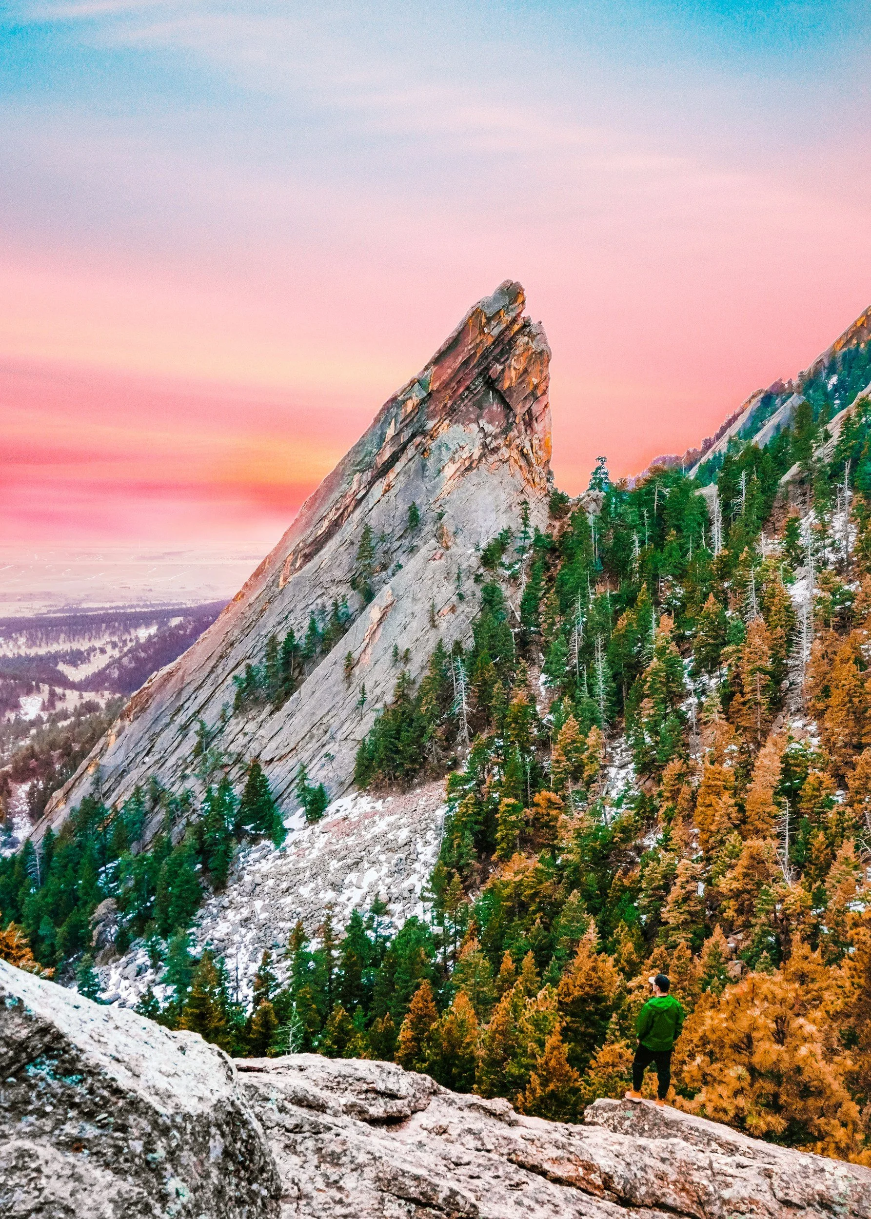A person in a green jacket and black pants standing on a rocky ledge, looking at a large, pointed granite mountain surrounded by a forest of pine trees, with a colorful sunset sky in the background.
