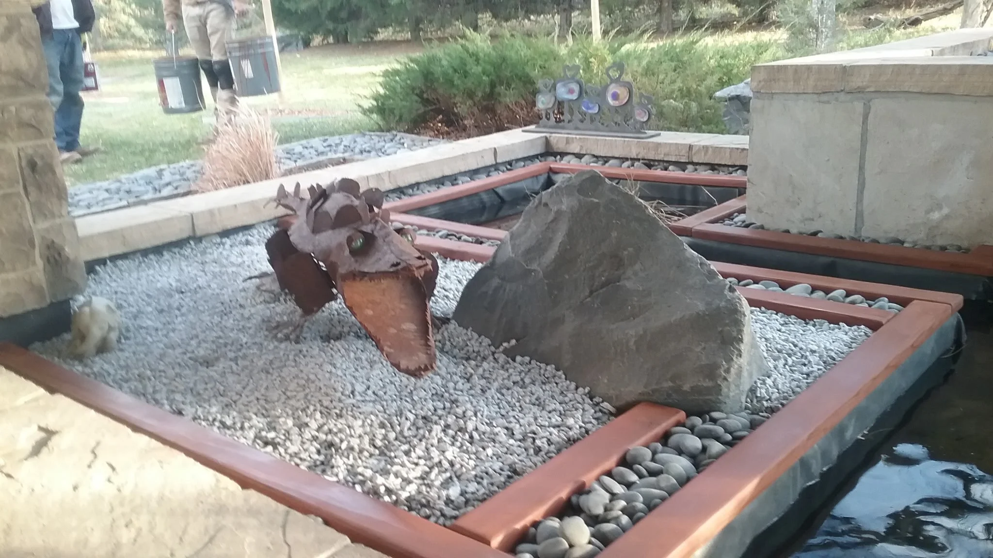Decorative pond with rocks, a metal reindeer sculpture, and a river stone border, with people in the background.