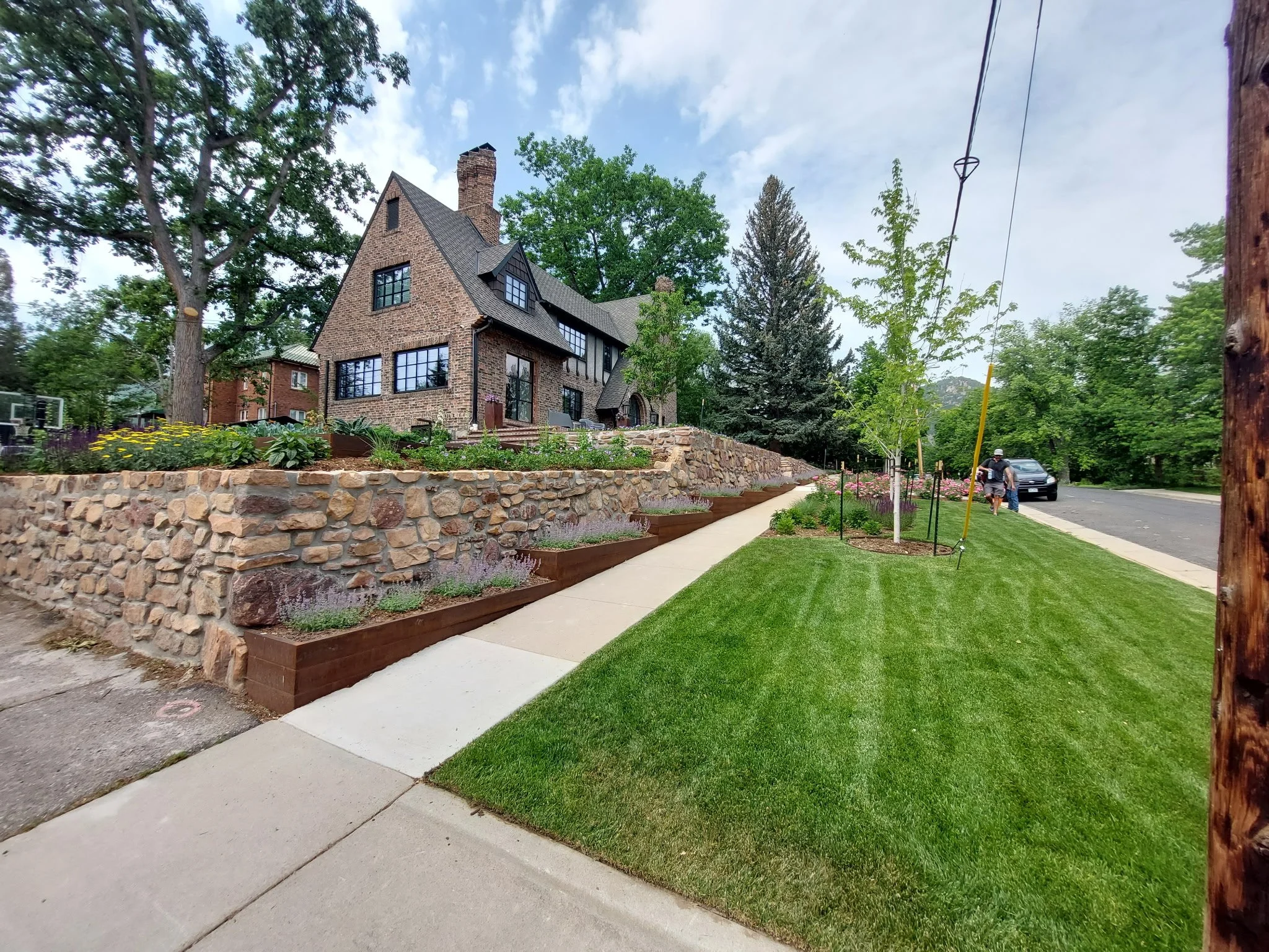 A stone house with black window frames on a grassy hill, landscaped yard with flower beds, sidewalk, and street with parked cars, trees, and pedestrians.