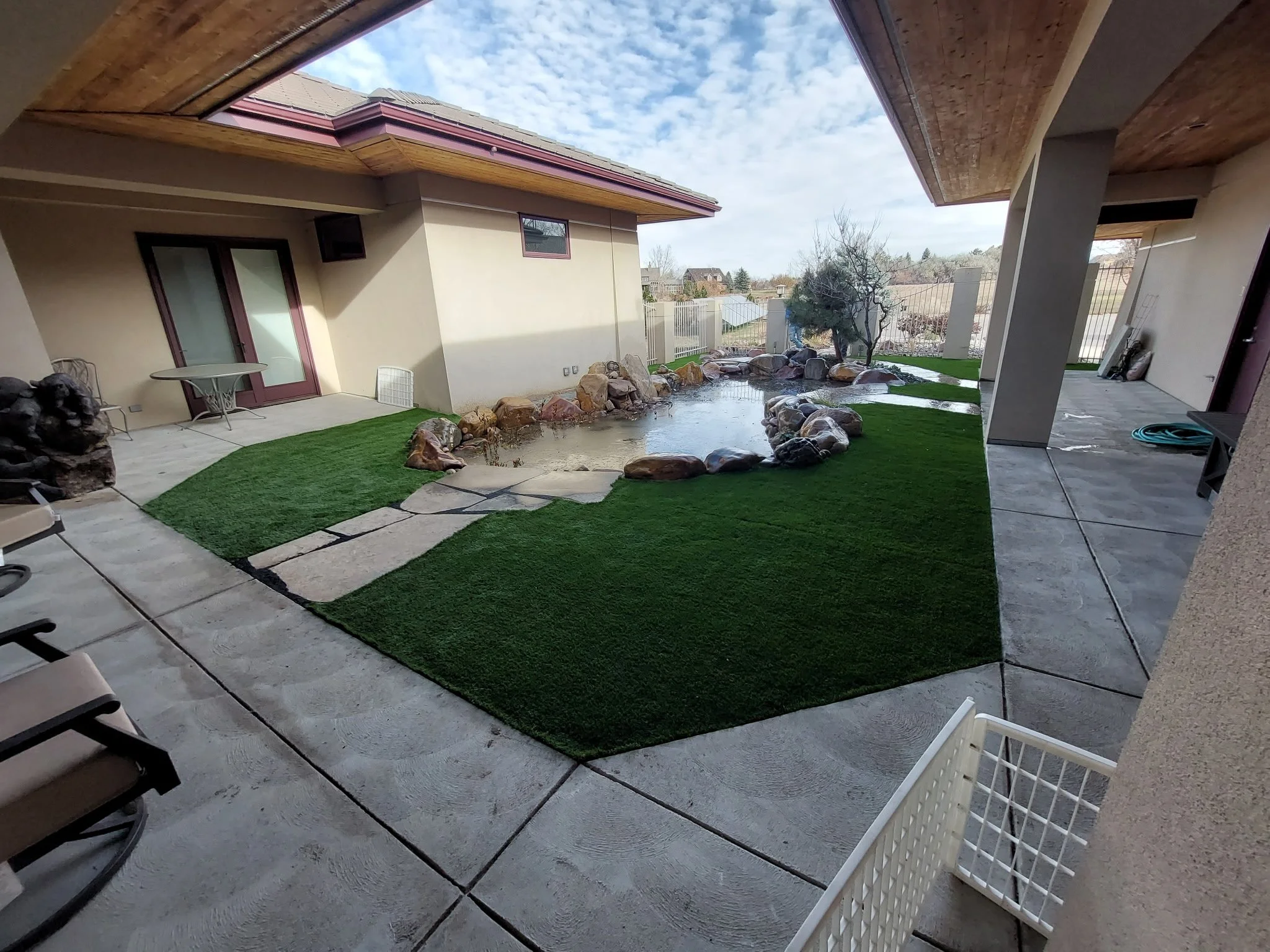 A backyard patio with a small pond surrounded by rocks, artificial grass, and a tree. There are chairs, a table, and a garden hose.