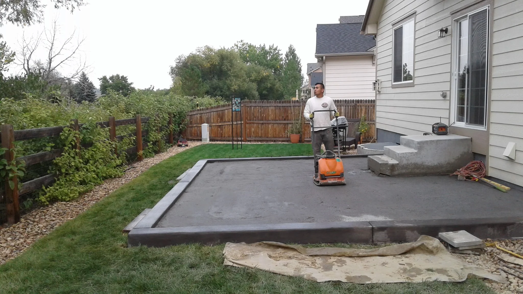 A man using a plate compactor to level concrete on a backyard patio area, with new concrete steps and surrounding grass and garden fencing.