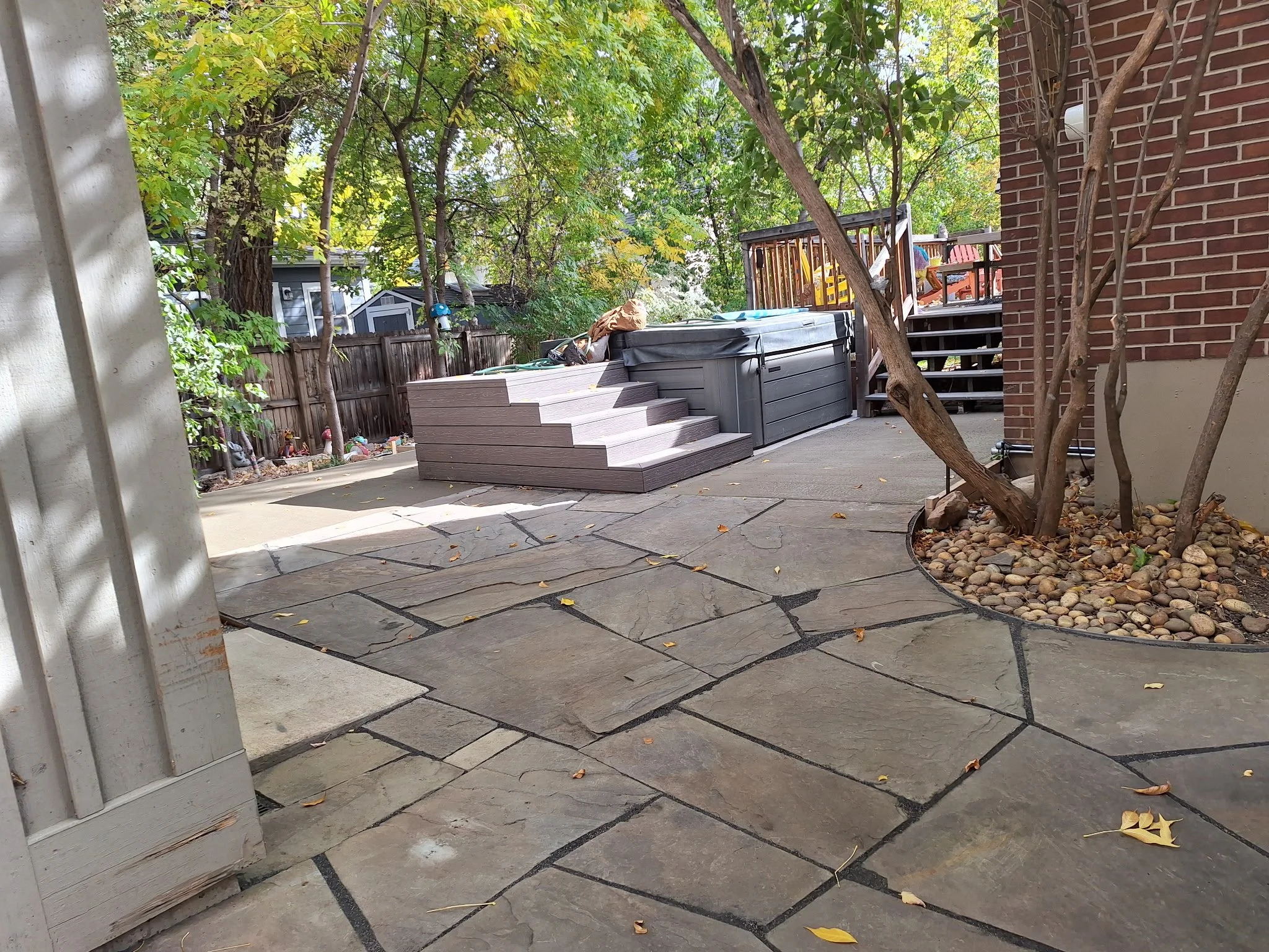 Decorative stone patio with tan, gray, and brown stones, a tree with a circular mulch bed, a staircase leading to a deck, and a hot tub, in a backyard with a fence and trees.