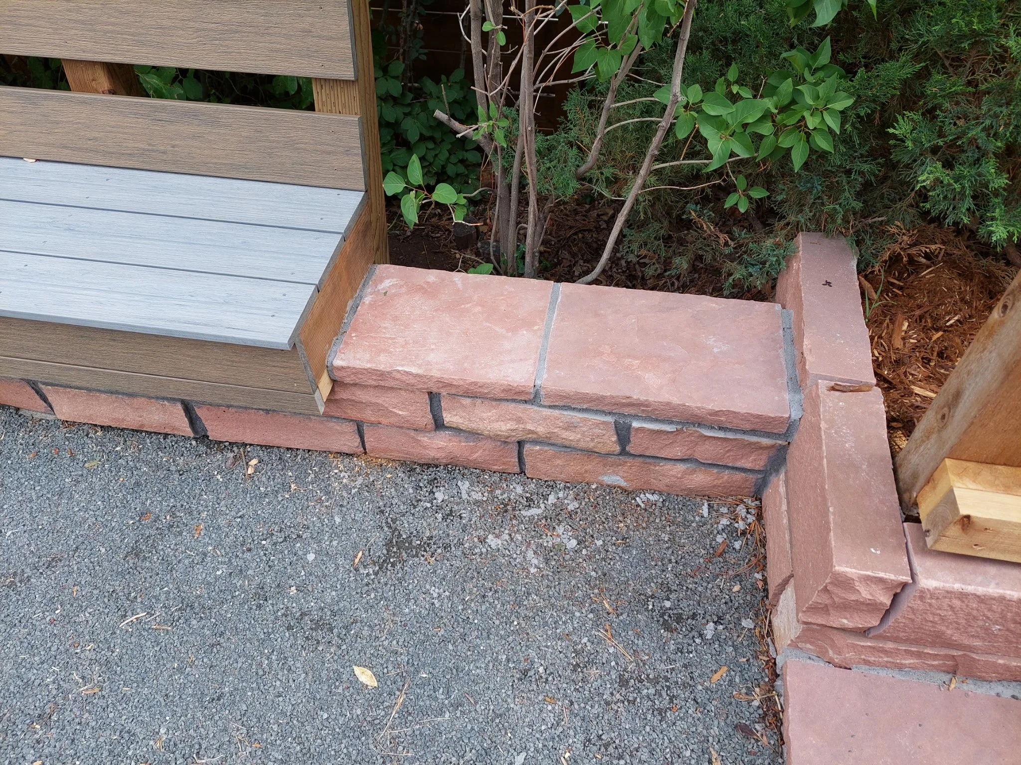 Corner of a constructed brick border with a wooden fence and green bushes behind it. The brick border has a step with pinkish bricks on top of a row of rectangular bricks. The ground next to it is gravel.