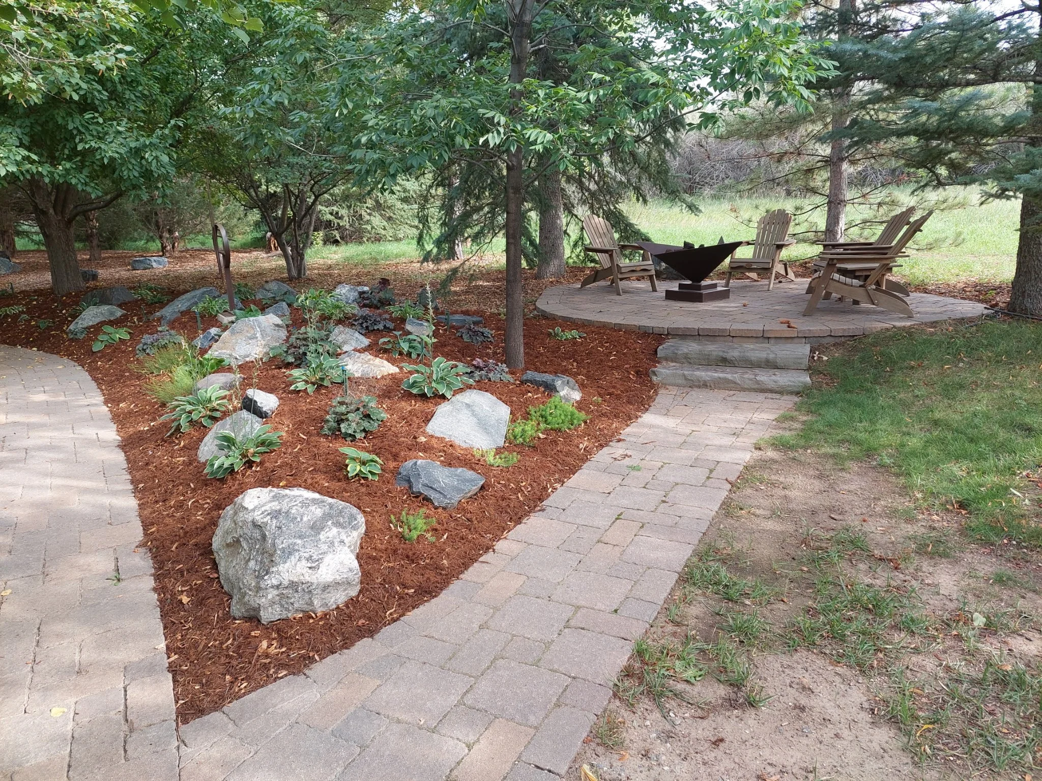 Outdoor seating area with Adirondack chairs around a fire pit on a circular stone patio, surrounded by trees and a garden bed with rocks and plants, with a brick pathway leading to it.