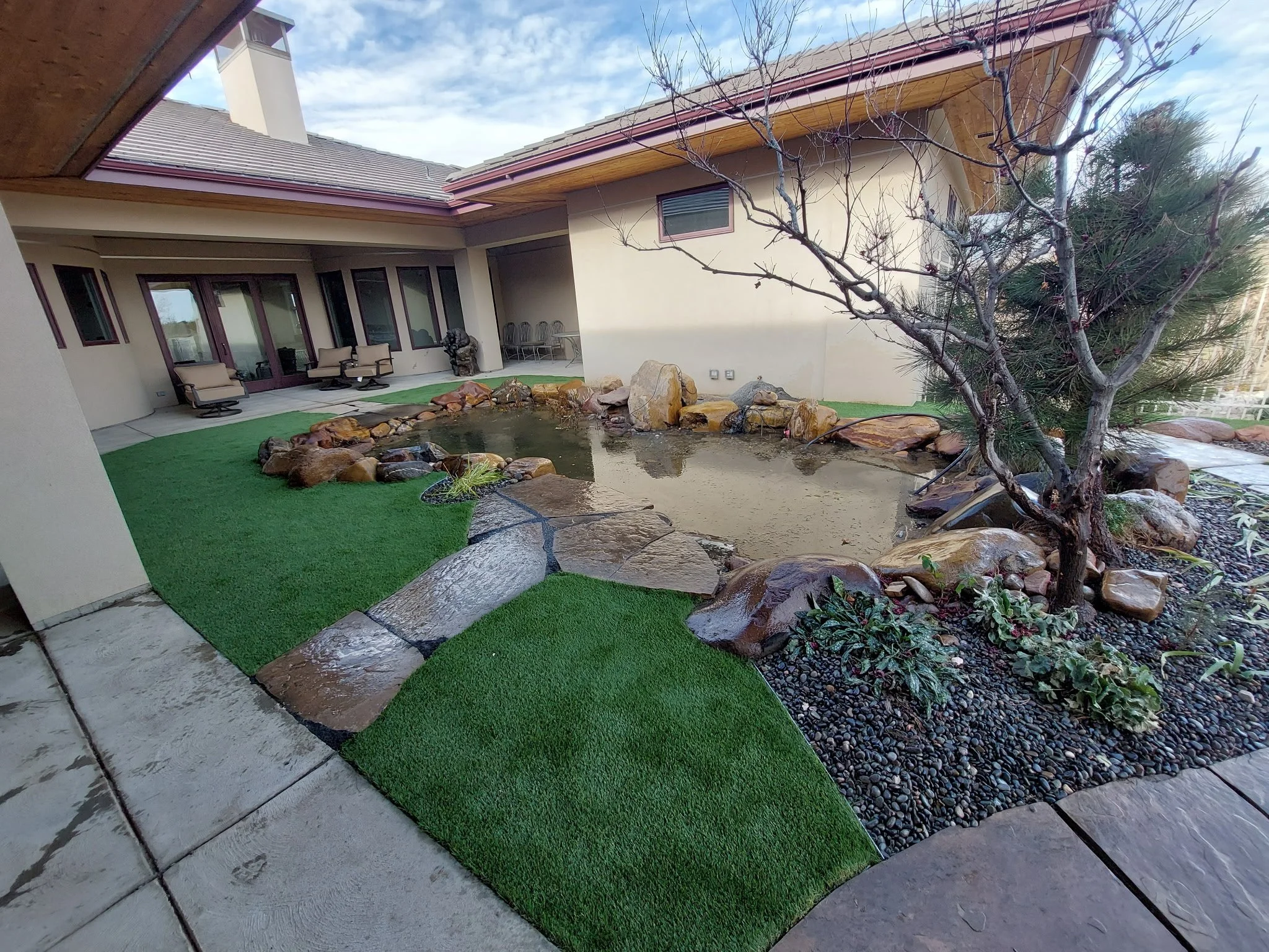 Backyard with artificial grass, a pond with rocks, a leafless tree, and outdoor seating area near a house.