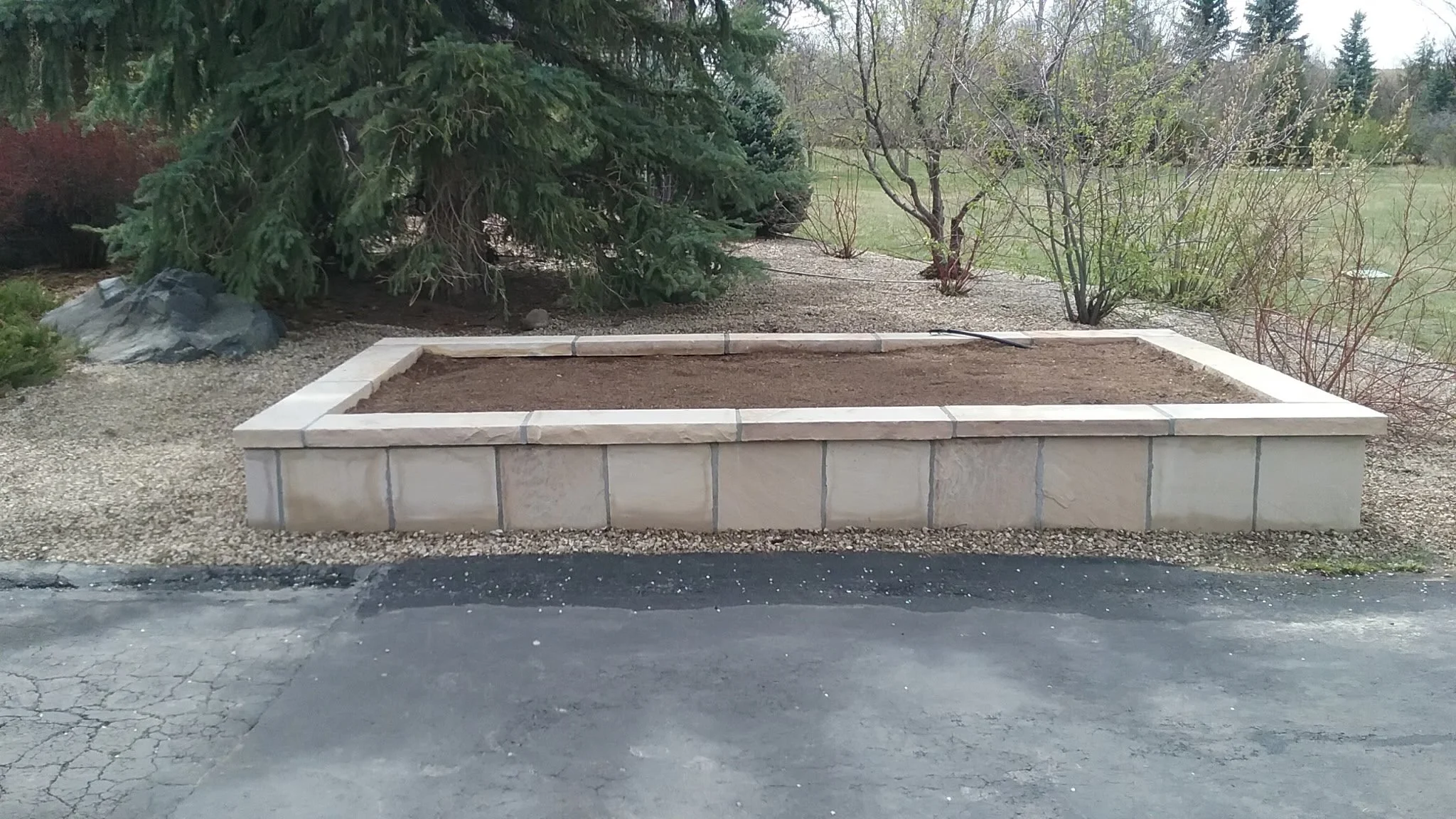 A rectangular raised garden bed with beige stone sides filled with soil, situated on a paved surface with a background of trees and bushes.