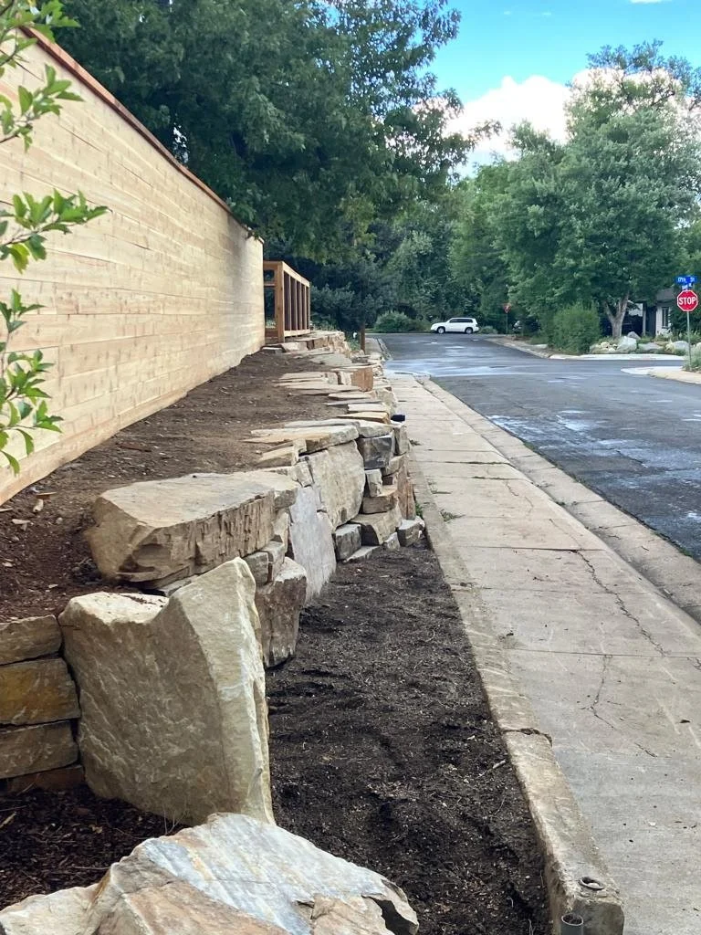 Sidewalk and a partially constructed stone retaining wall beside a house with a wooden fence, with trees and a parked car in the background.