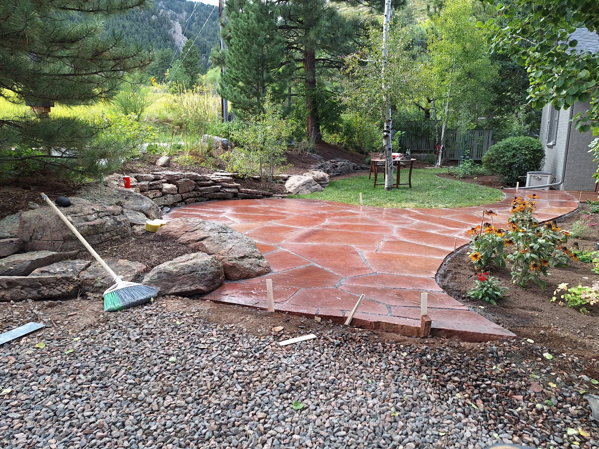 A backyard garden with a newly paved stone patio, surrounded by trees, plants, and garden tools, with a gray house visible on the right.