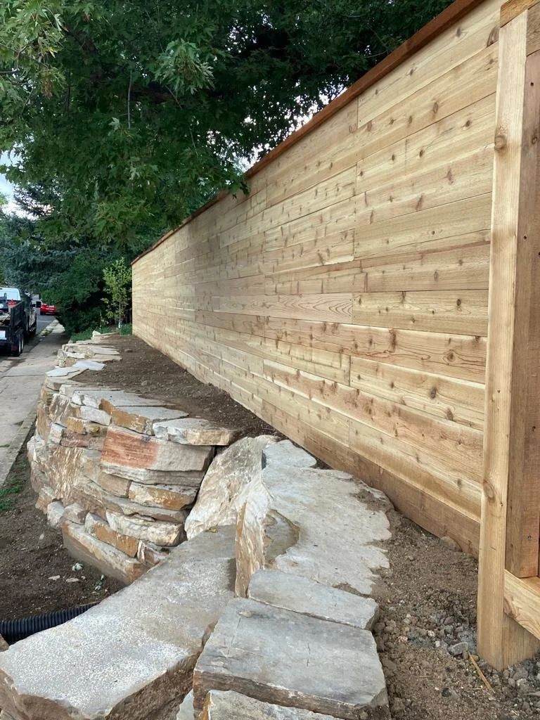 A newly built wooden fence runs along the right side of the image, with a stone pathway and a stone retaining wall with irregularly shaped rocks in the foreground. There are trees and parked cars visible in the background.