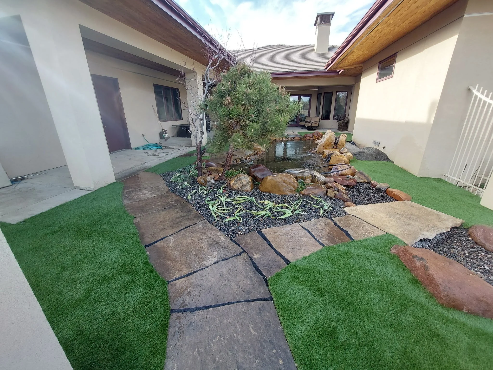 Backyard with artificial green grass, stone pathway, small pond with rocks, and trees in a residential area.