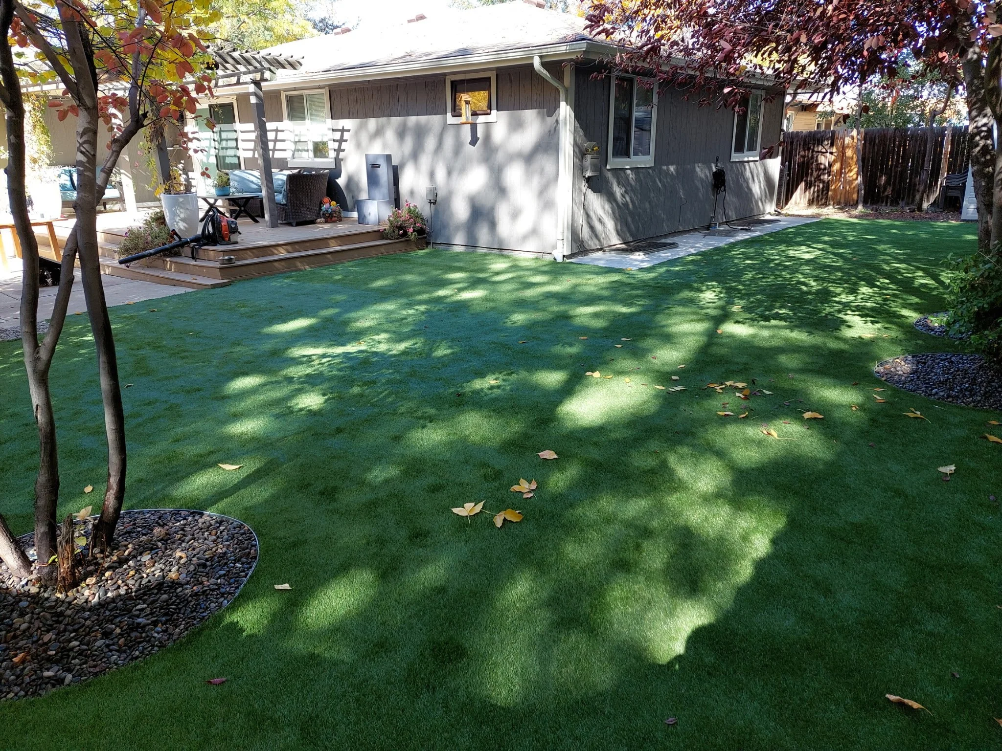 Backyard with a well-maintained lawn, trees with red leaves, a small deck, and a gray house in the background.