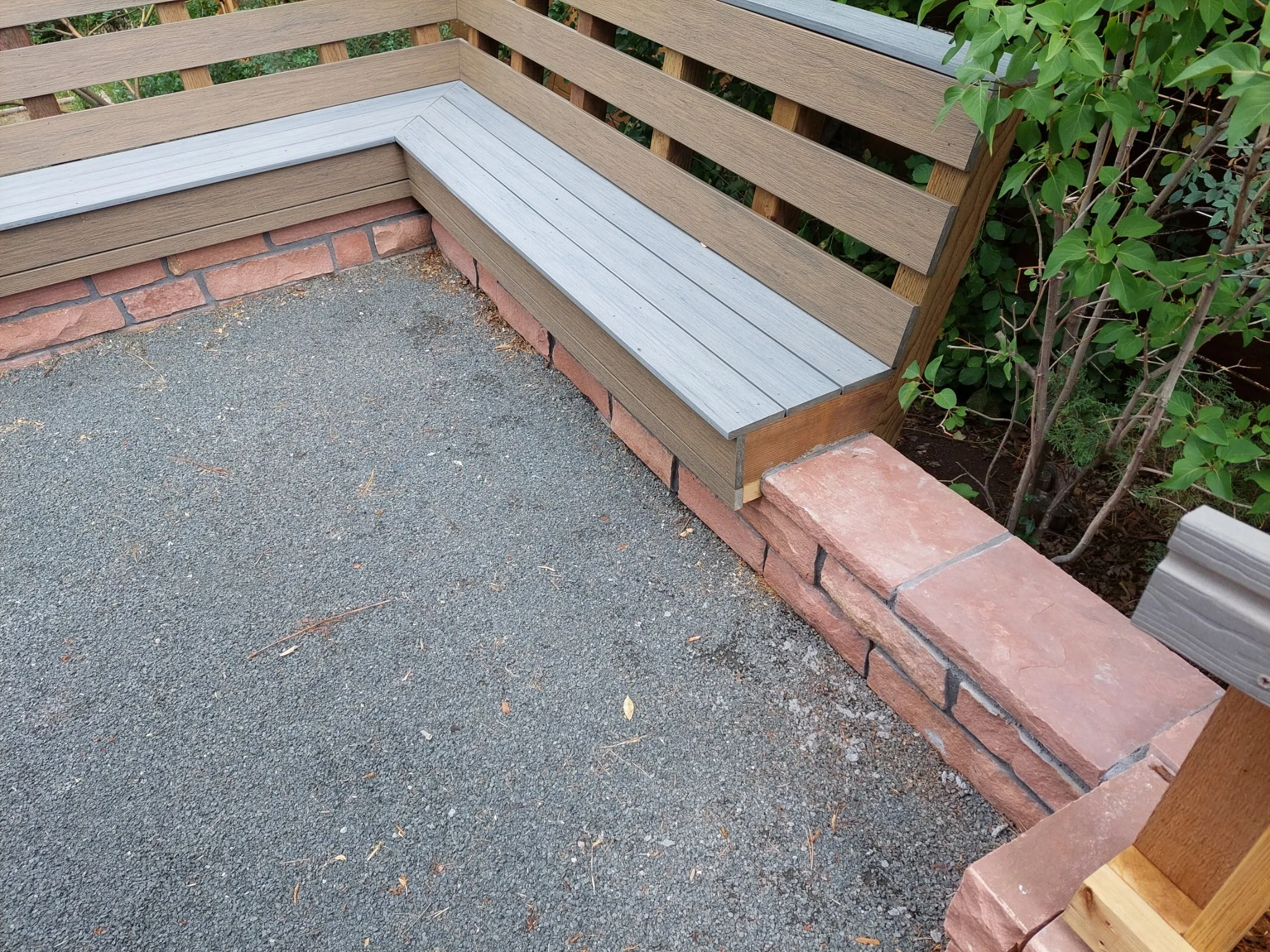 A corner outdoor space featuring a gray wood bench with a backrest, built on a raised brick platform. The ground is paved with textured asphalt, and green foliage is visible behind the wooden railing.