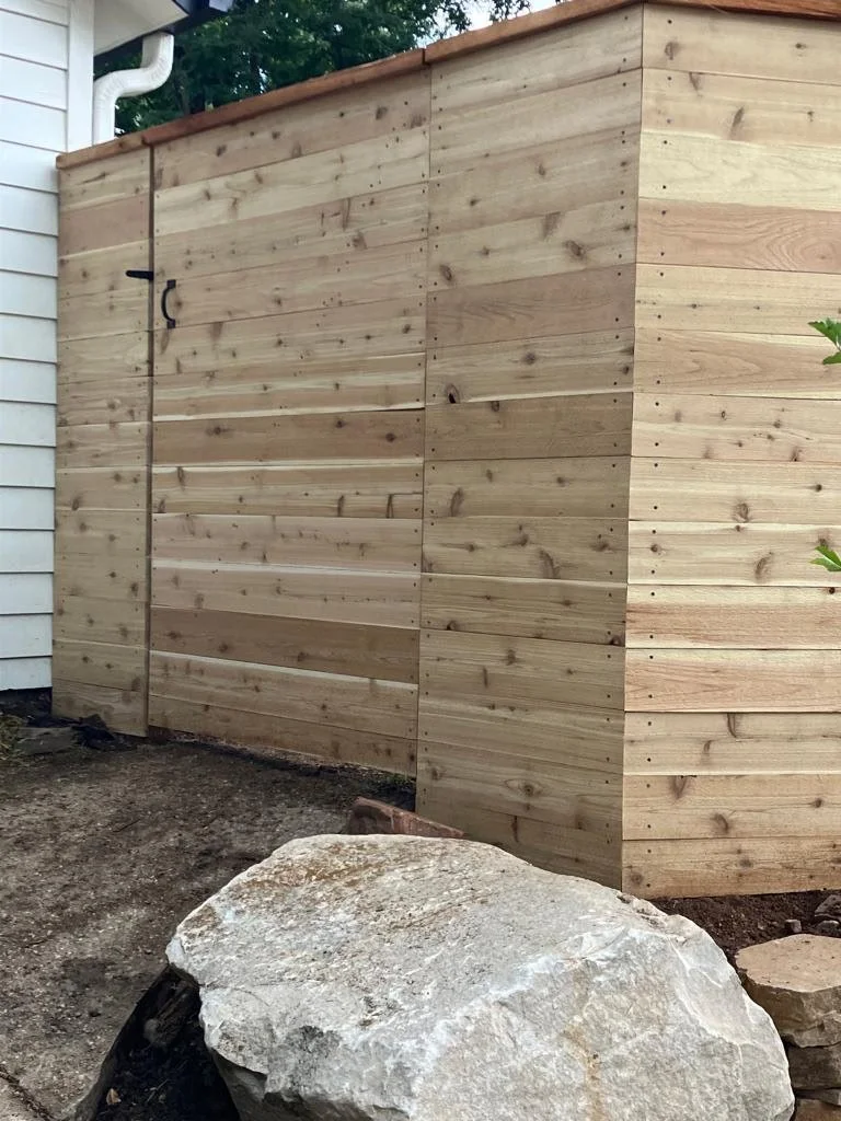 A wooden privacy fence next to a house with white siding. There is a large rock in the foreground and some dirt around it.