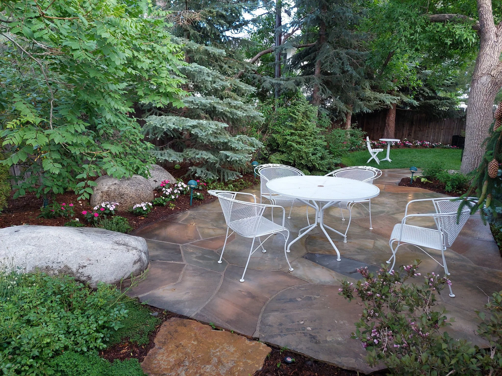 A backyard patio with white metal chairs and a table, surrounded by greenery, trees, and flowering plants.