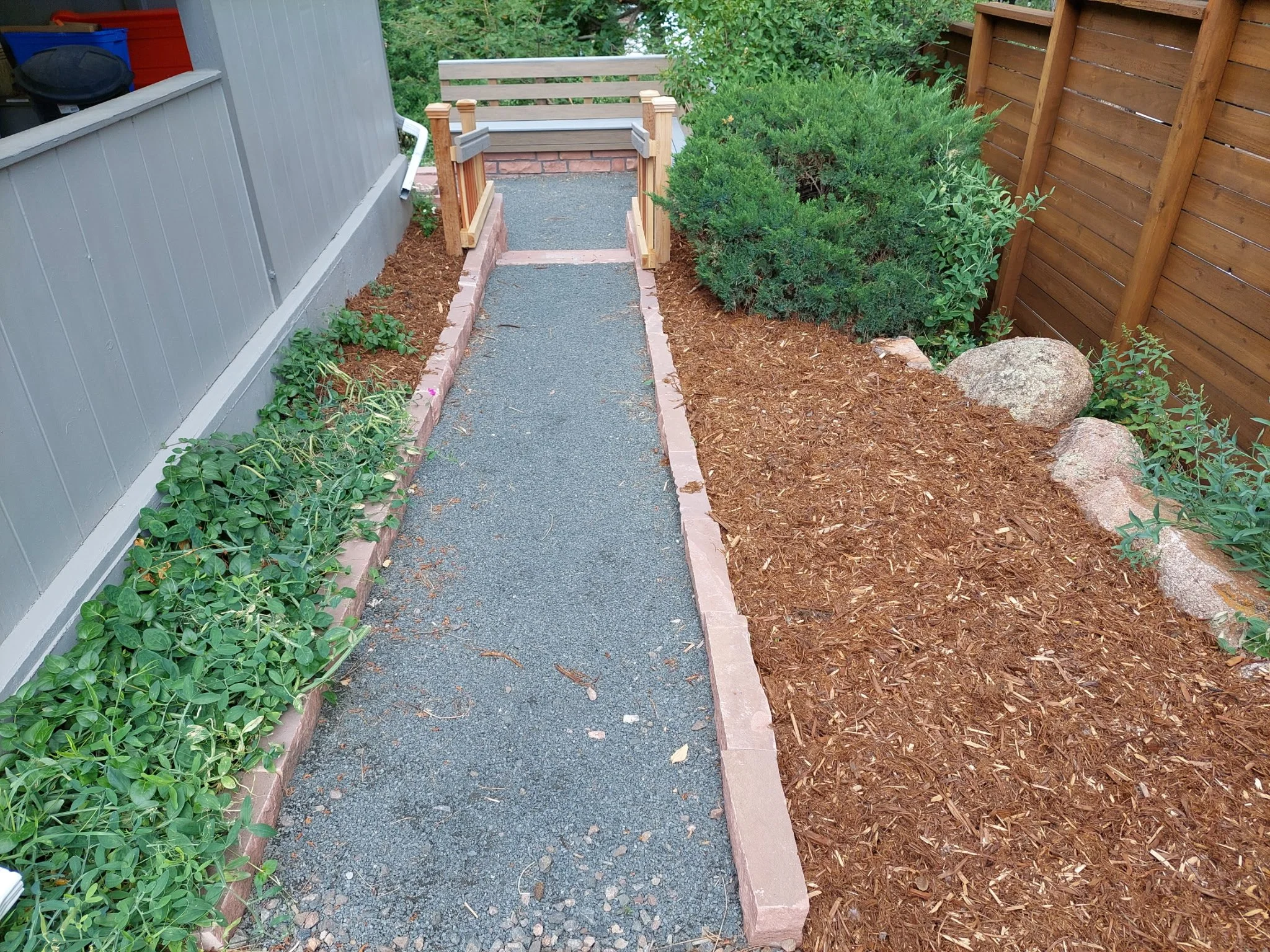 A small outdoor pathway with a gravel surface, bordered by brick edging, leading to a wooden ramp with railings. On the left side, there's a grey house wall with some green plants and a black vent. On the right, there's a brown mulch bed with rocks a