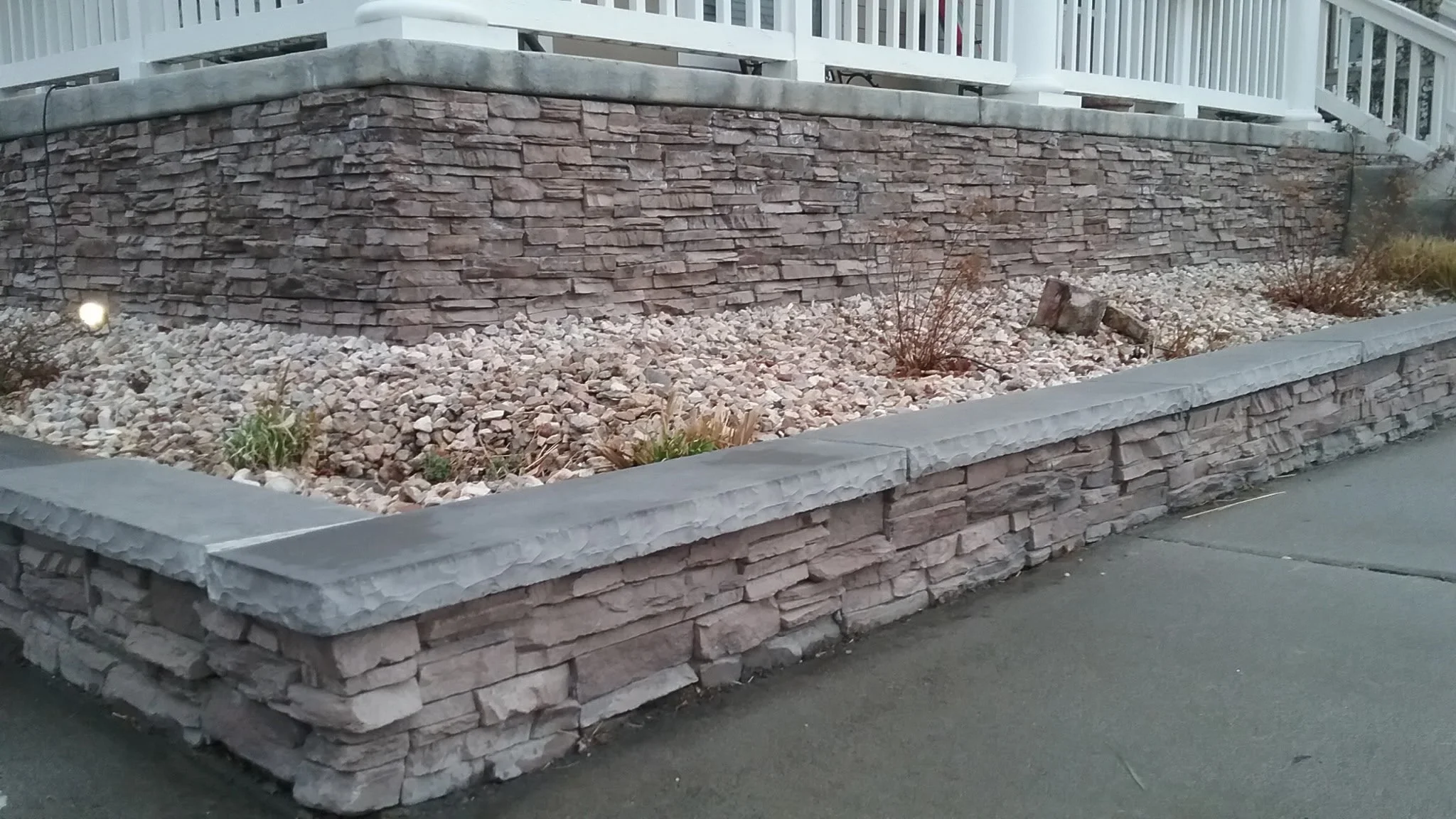 Stone retaining wall with a landscaped area filled with small rocks and sparse plants in front of a sidewalk.