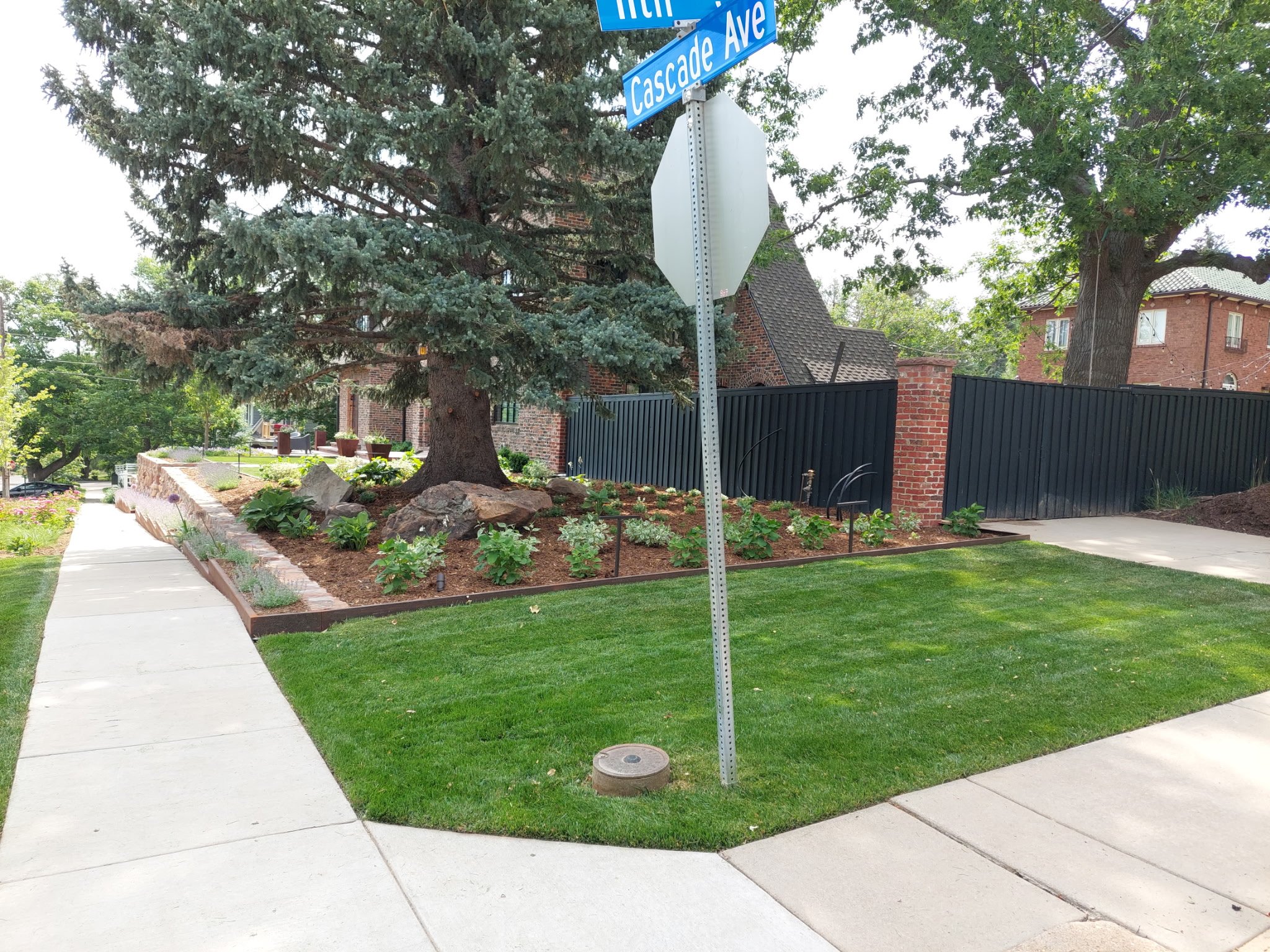 A residential neighborhood corner with a sidewalk, a well-kept lawn, and a garden with trees and plants. Blue street signs indicate the intersection of Highland Drive and Cascade Avenue.