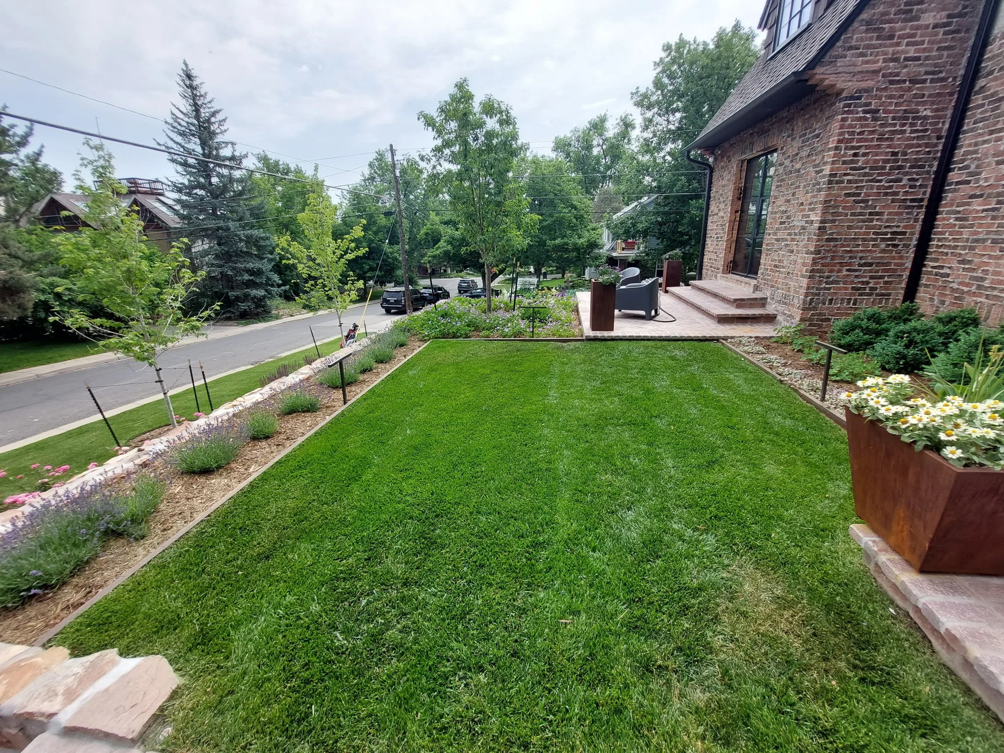 A well-maintained front yard with a lush green lawn, flower beds along the sidewalk, small trees, and a patio with outdoor furniture next to a brick house.