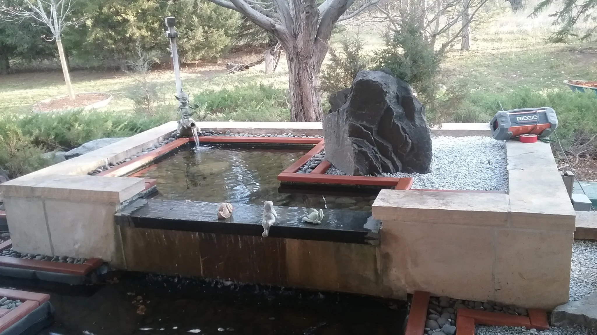 A backyard water feature with a stone and concrete structure, small waterfalls, a large dark rock, small decorative frogs, and a portable speaker on the right side.