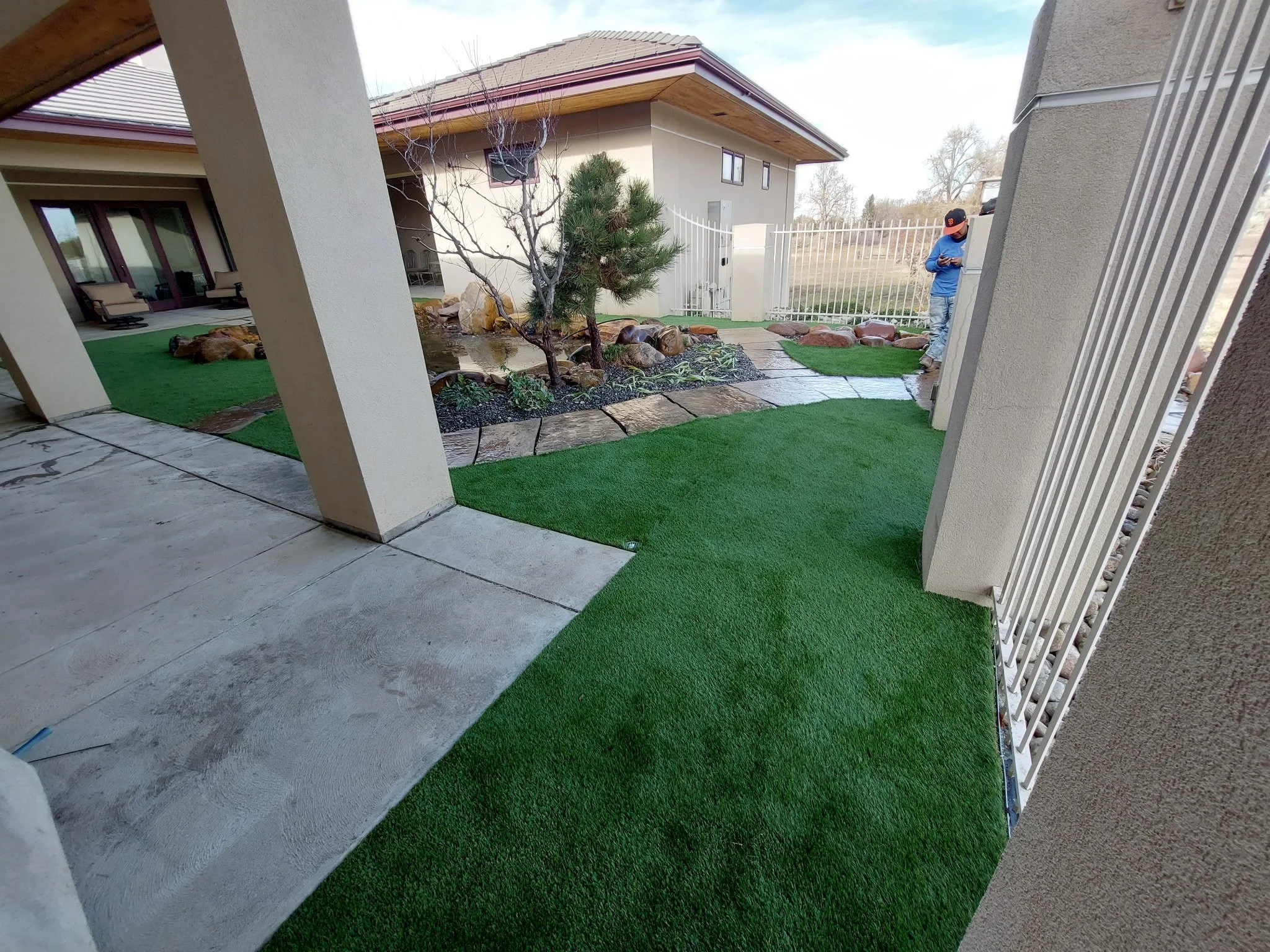 View of a backyard patio with artificial grass, stone walkway, small trees, and two people standing by the fence.