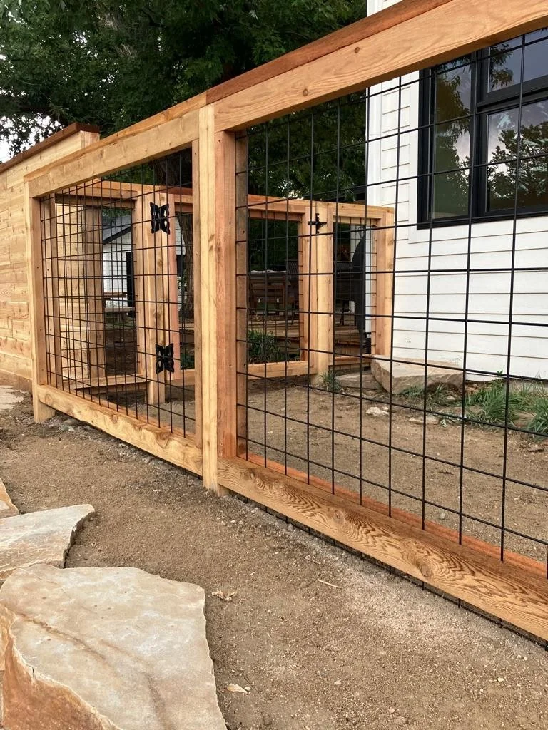 Wooden and wire fence structure next to a white house with black window frames, with trees and rocks nearby.
