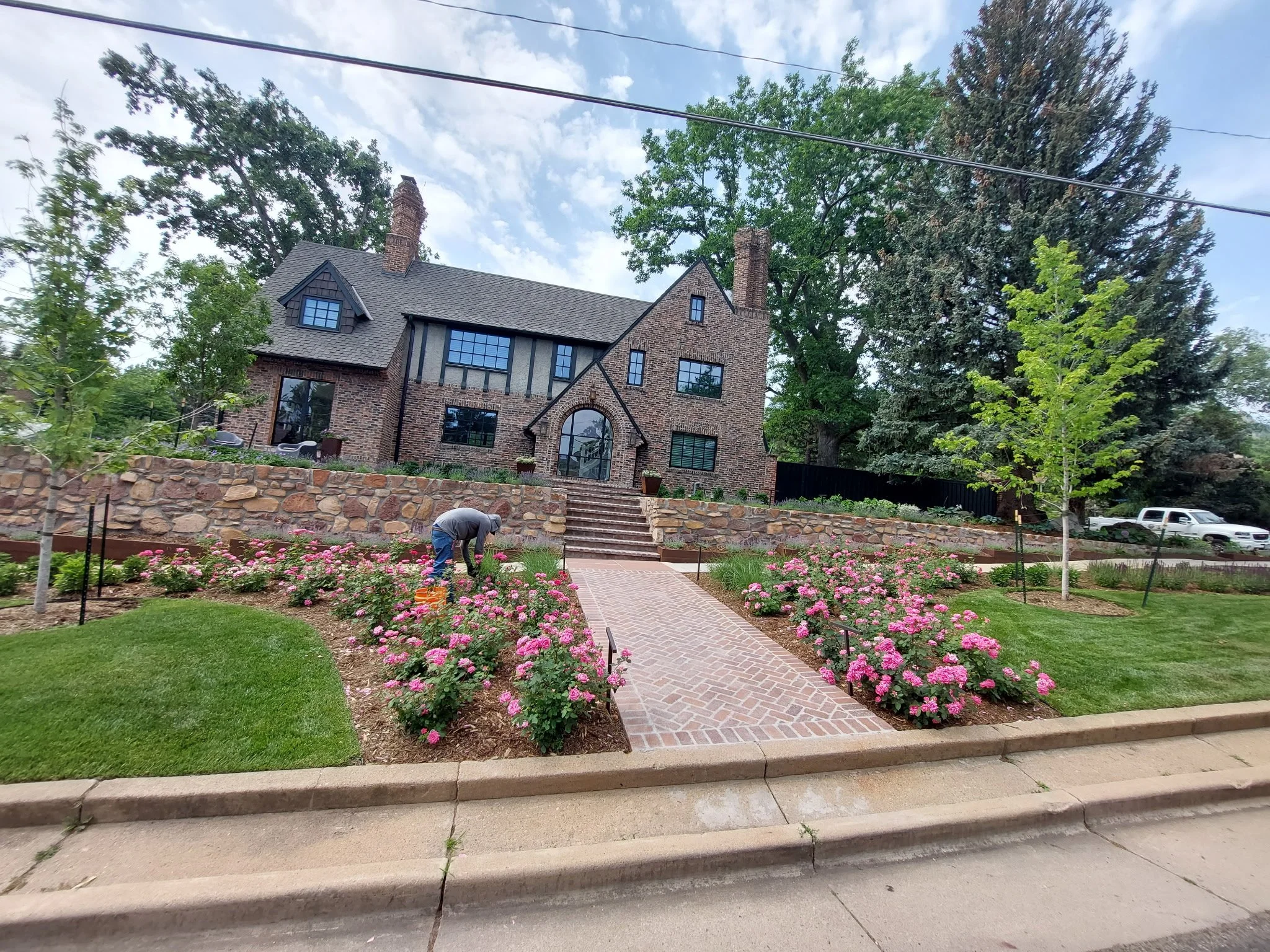 A person tending to pink flowering plants in a landscaped yard in front of a large brick house.