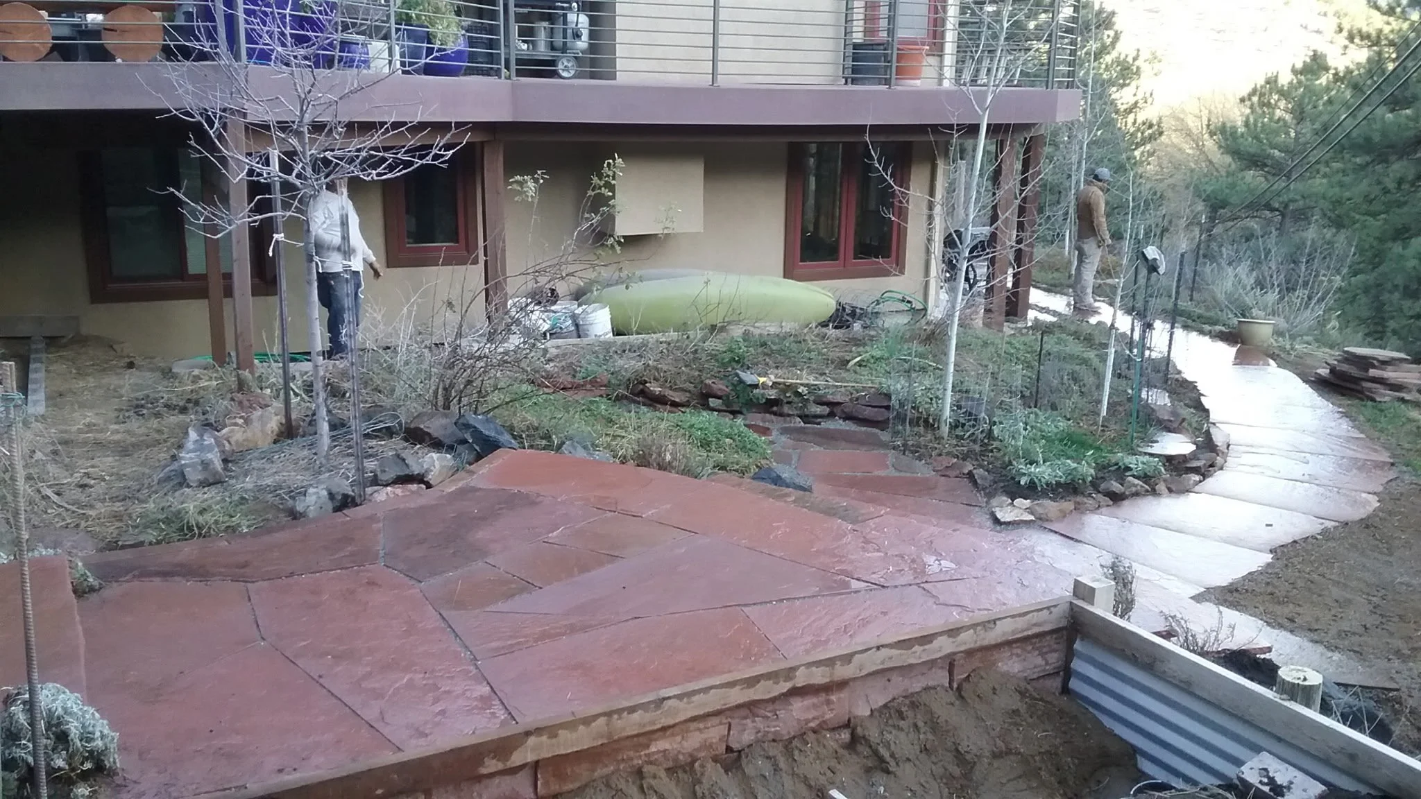Backyard landscape with new stone pathway, leafless trees, and garden beds under construction, in front of a house with beige walls and red window frames.