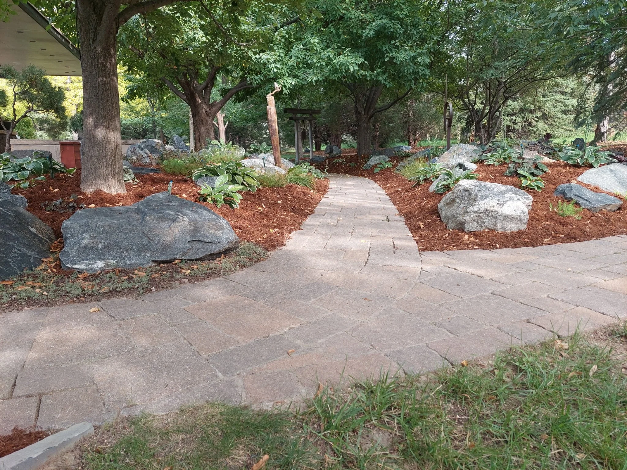 A winding brick pathway through a landscaped garden with large rocks, mulch, green plants, and trees.