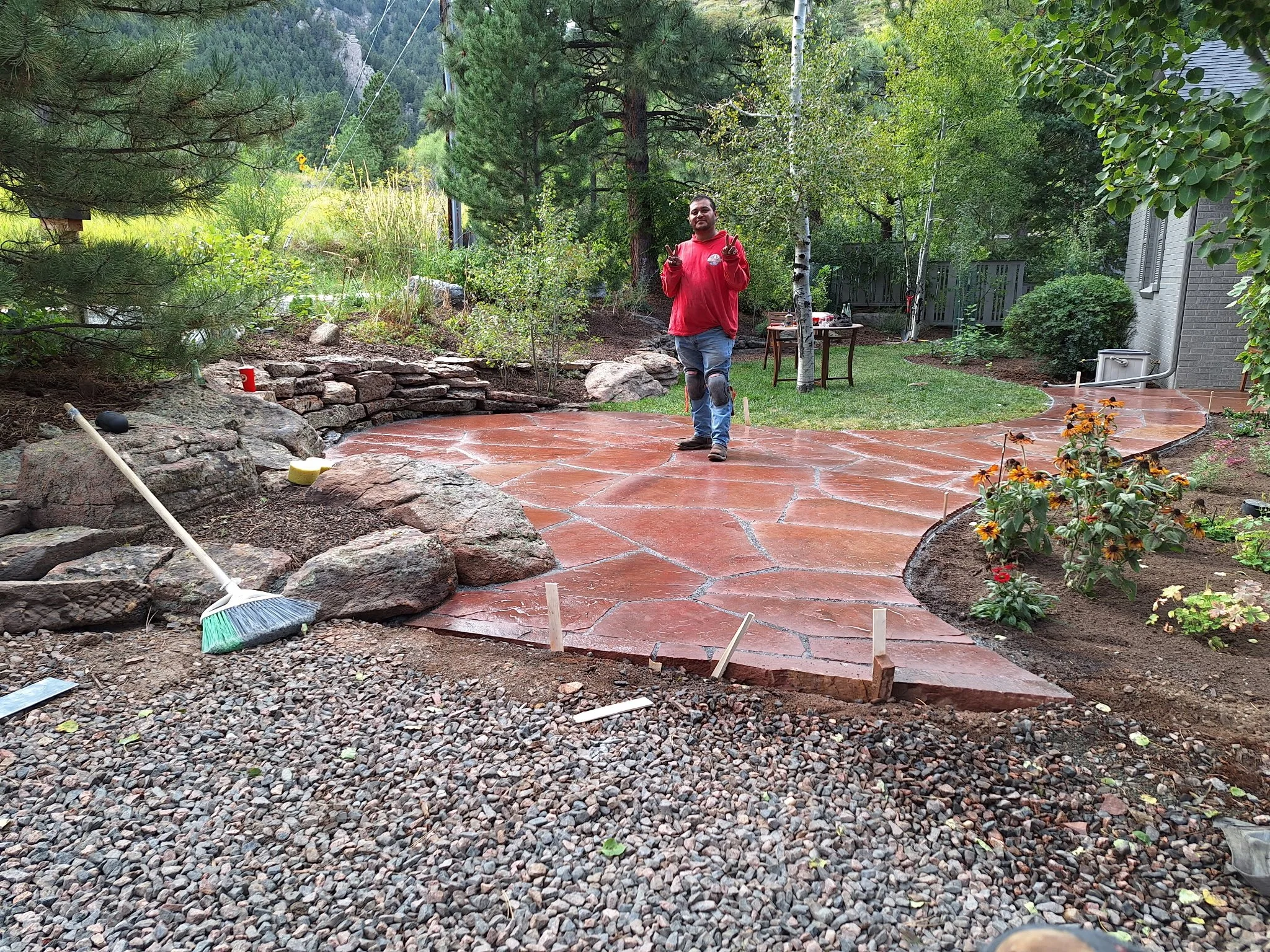 A man in a red hoodie and jeans with patched knees is walking on a newly paved red concrete patio in a backyard garden. There are large rocks and gardening tools, including a broom and a yellow scrubbing pad, on the ground near the patio. The area is